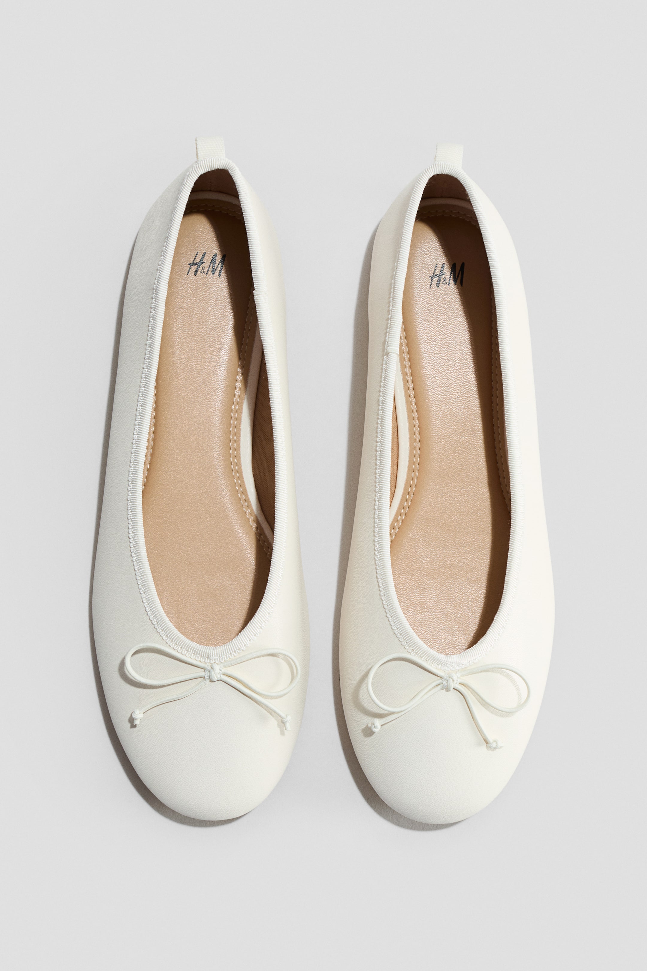 Visa större bild: Ett par off-white ballerinaskor ligger platt, sedda ovanifrån. Varje sko har en smal, elastisk kant runt öppningen och visar en dekorativ rosett vid främre tån. De ljusbruna innersulorna, märkta med "H&M"-logotypen, är synliga inuti.