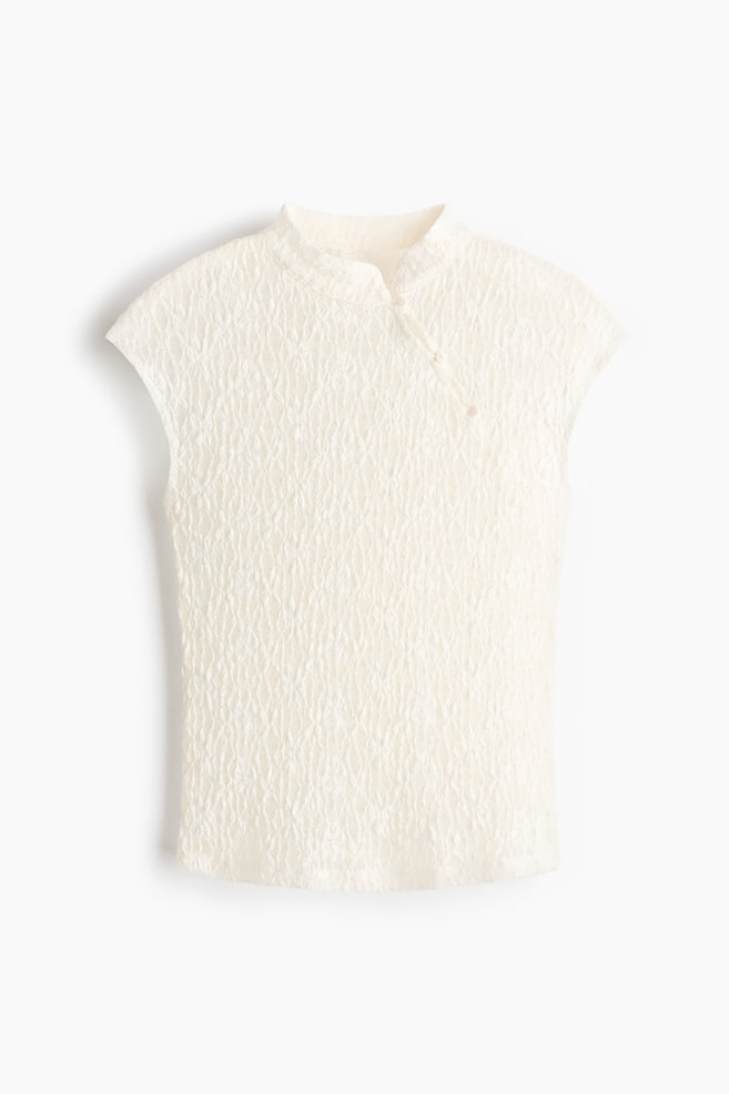 Un top de color blanco roto con textura arrugada, cuello alto, cierre frontal asimétrico con dos botones de nudo y mangas tipo capa. - 1