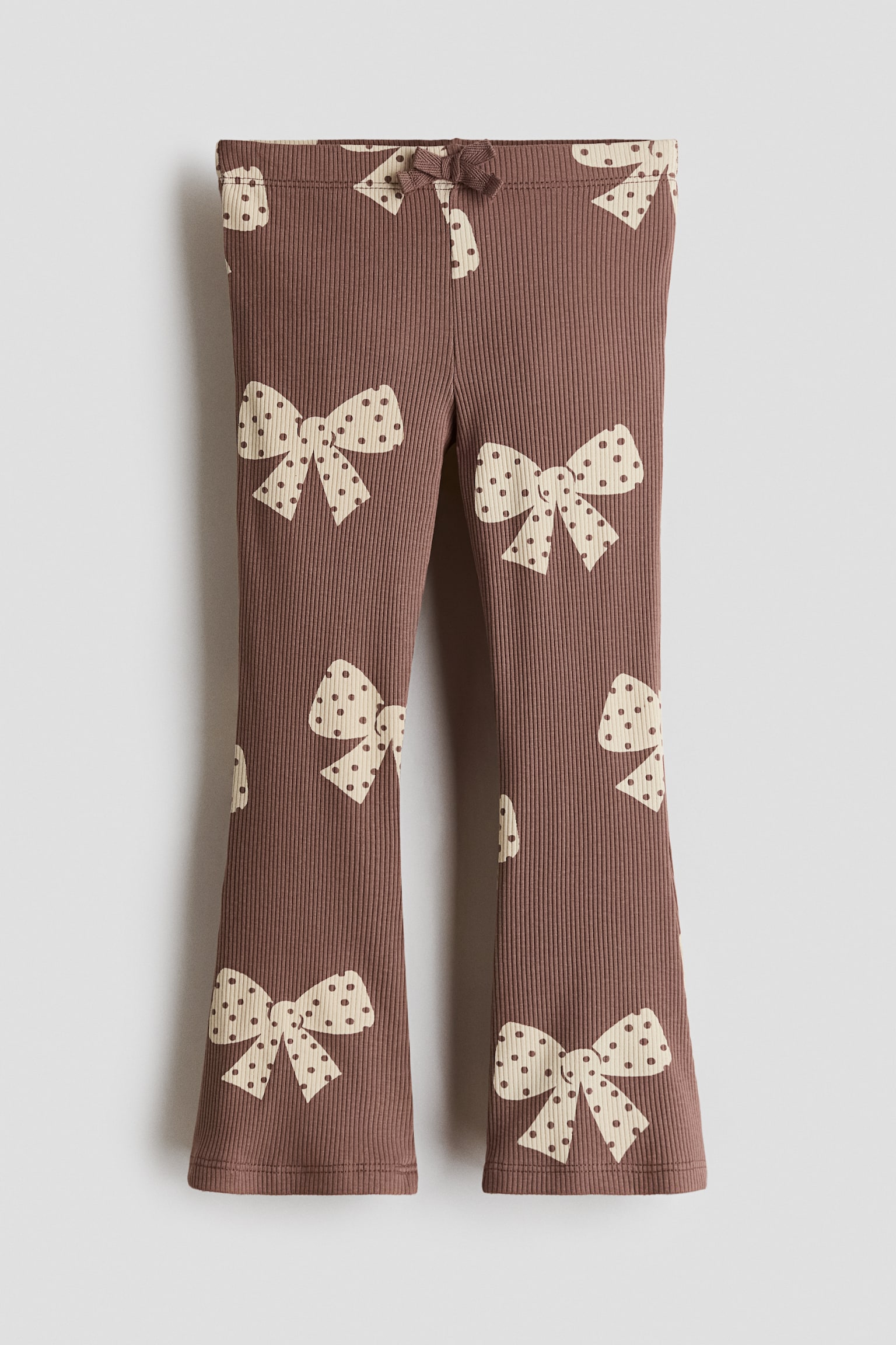 These brown ribbed cotton jersey leggings are shown from the front, featuring an all-over pattern of cream-coloured, polka-dotted bows. The design includes an elasticised waistband with a small decorative bow at the centre and flared legs.