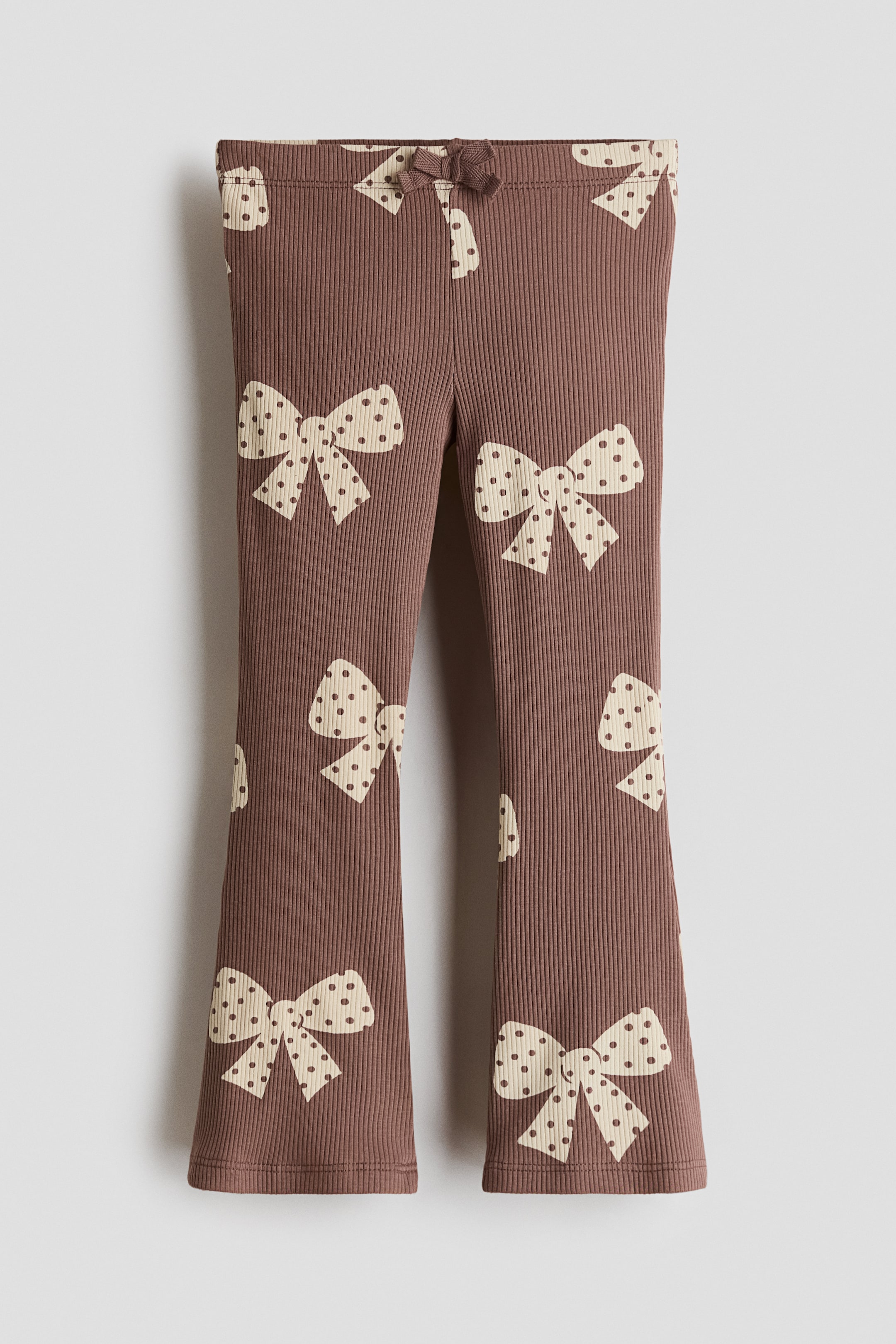 Xem ảnh lớn hơn: Những chiếc quần legging jersey cotton gân màu nâu này được chụp từ phía trước, nổi bật với họa tiết nơ chấm bi màu kem phủ toàn bộ bề mặt. Thiết kế gồm cạp chun với một chiếc nơ trang trí nhỏ ở giữa và ống quần loe.