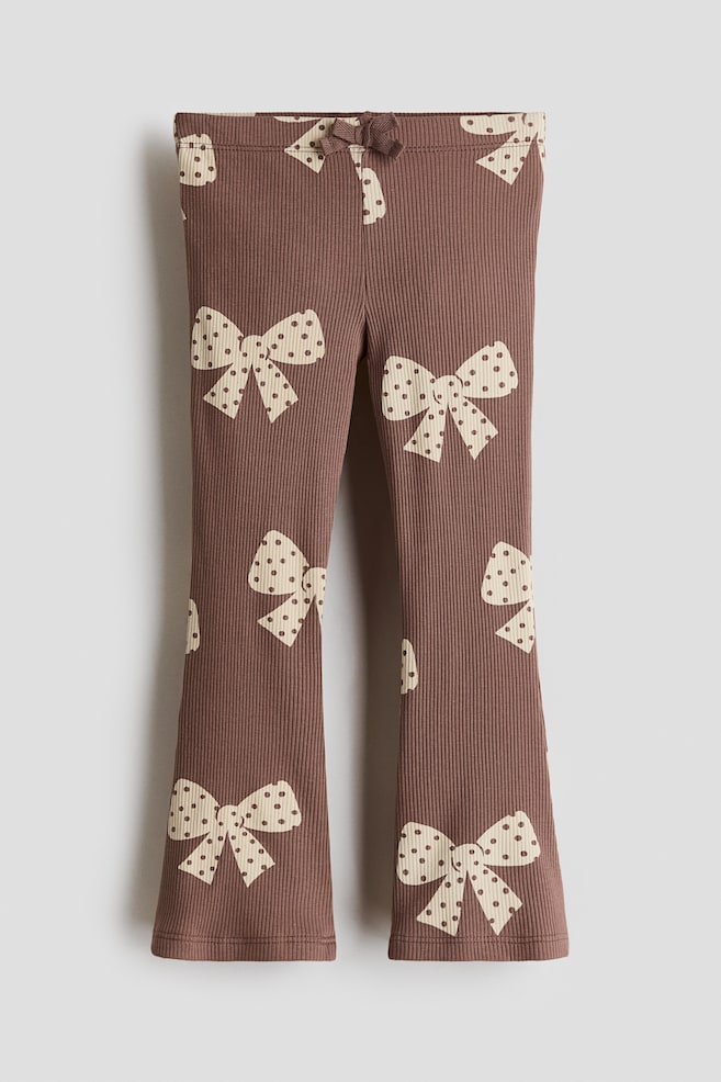 These brown ribbed cotton jersey leggings are shown from the front, featuring an all-over pattern of cream-coloured, polka-dotted bows. The design includes an elasticated waistband with a small decorative bow at the centre and flared legs. - 1