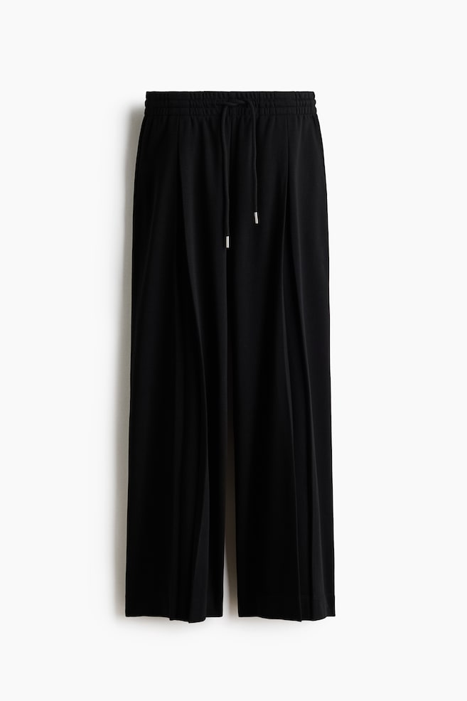 Black loose-fit trousers in lightweight sweatshirt fabric are featuring an elasticated drawstring waist with silver-tipped ties, and extra-long, wide legs, each displaying an inverted box pleat at the front. - 1