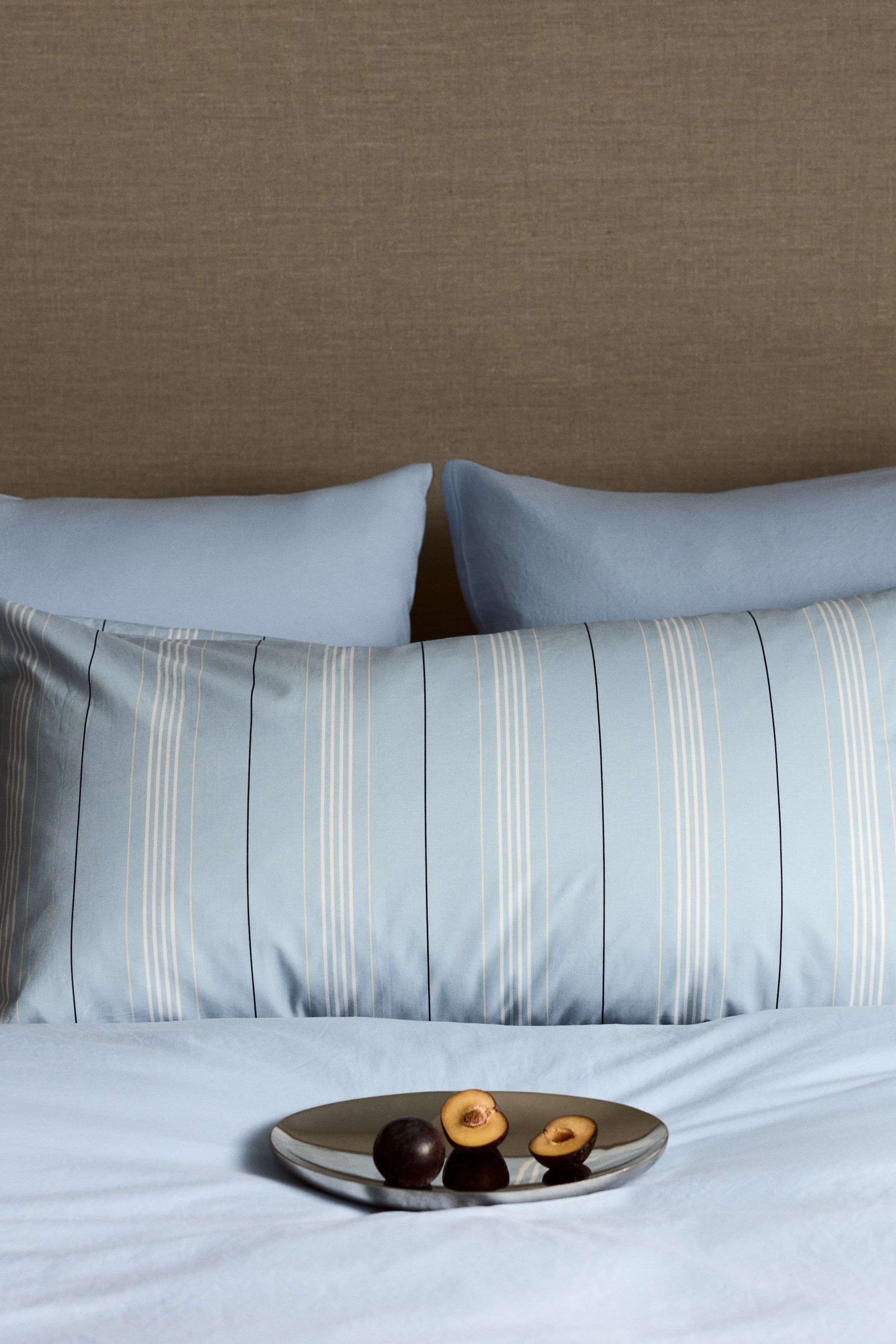View larger image: A light blue pillowcase features vertical stripes in white, beige, and thin black lines. It’s placed horizontally on a bed, layered over two solid light blue pillows and a coordinating light blue flat sheet.