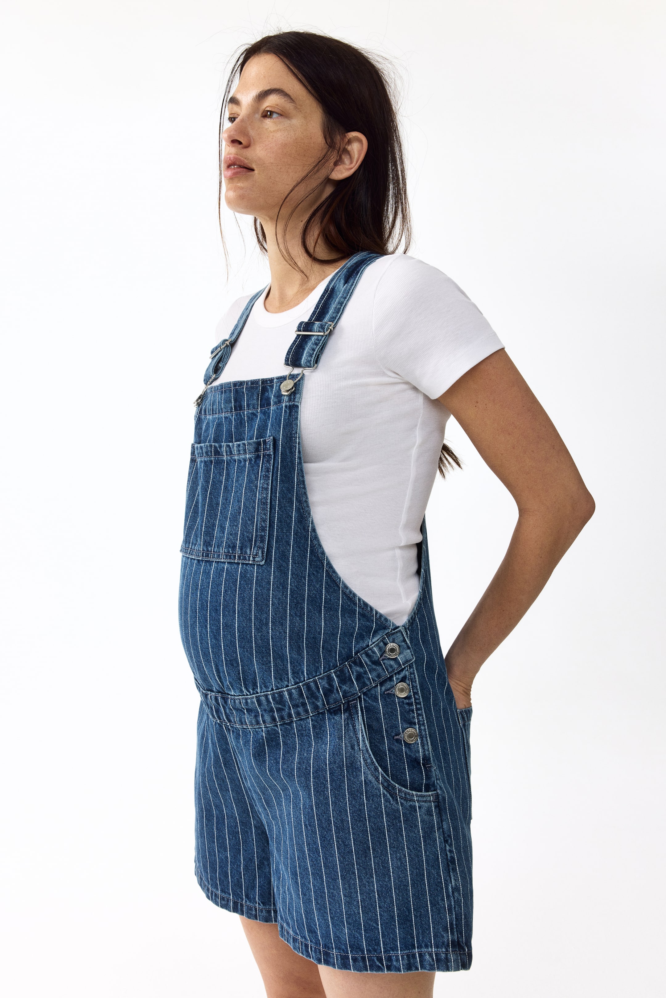 View larger image: Denim overall shorts are angled side-front, showcasing blue denim with white vertical pinstripes. They feature adjustable shoulder straps with metal buckles, a bib with a patch pocket, three metal buttons on each side of the waist, and side pockets, with a white t-shirt worn underneath.