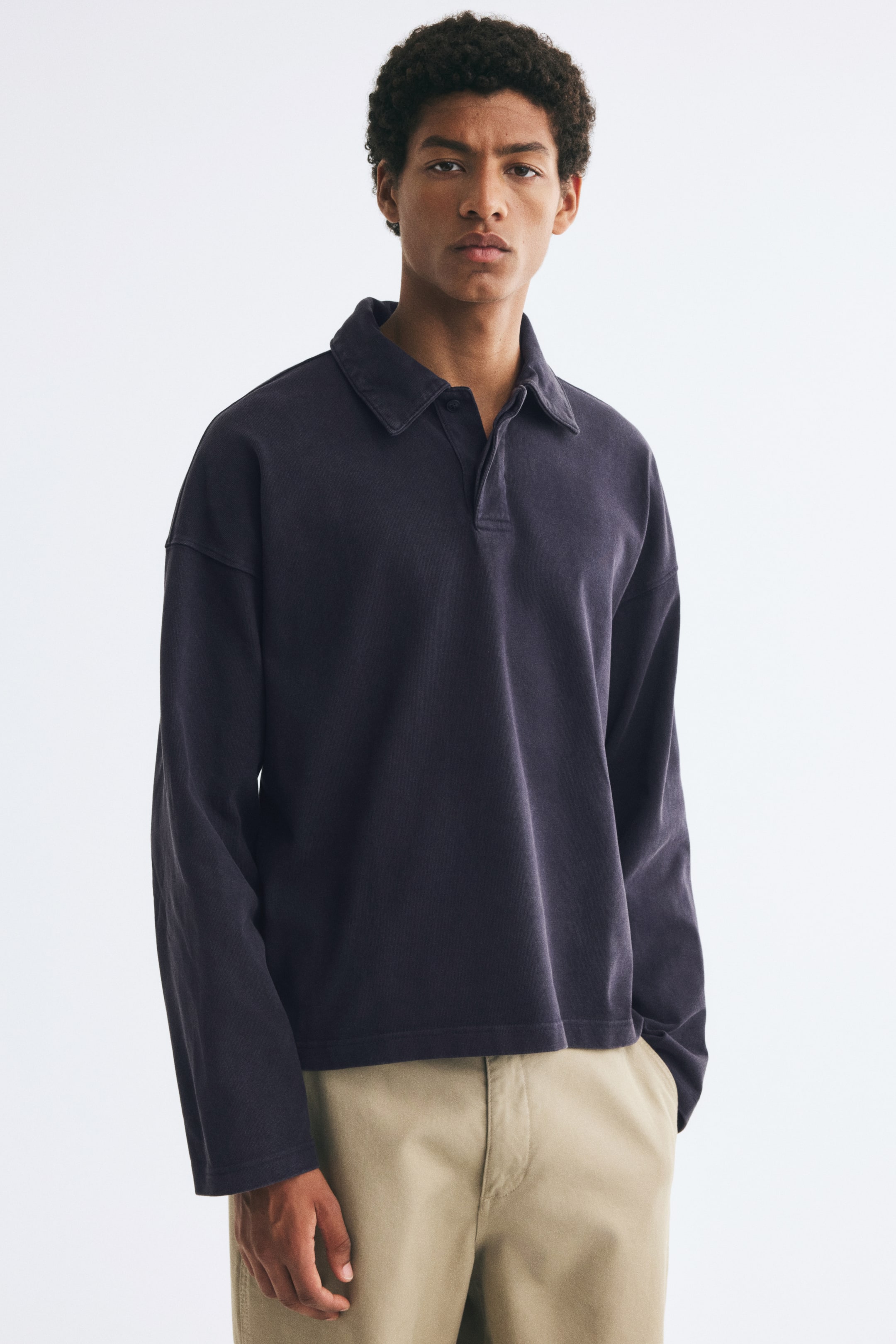 Ver imagen más grande: La modelo lleva una polo de manga larga en gris oscuro combinada con pantalones beige. La prenda presenta un corte oversize con hombros caídos, cuello clásico y bajo recto.