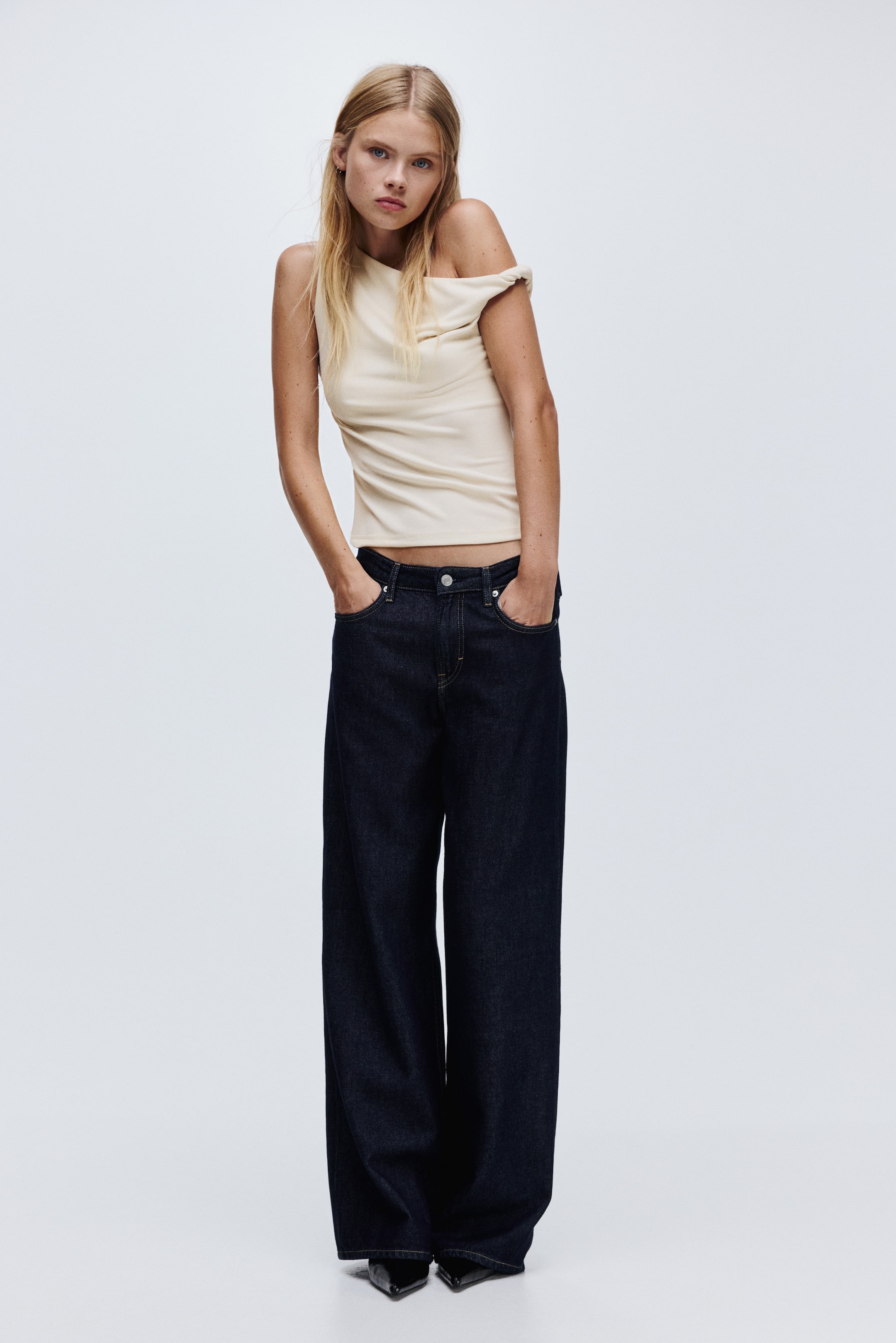 View larger image: A cream-colored, fitted top is presenting a one-shoulder design with a draped off-the-shoulder detail on the wearer's right side and a visible strap on the left. It is styled with dark wash, wide-leg jeans that are featuring front pockets with contrast stitching and are falling to a full length over the feet.