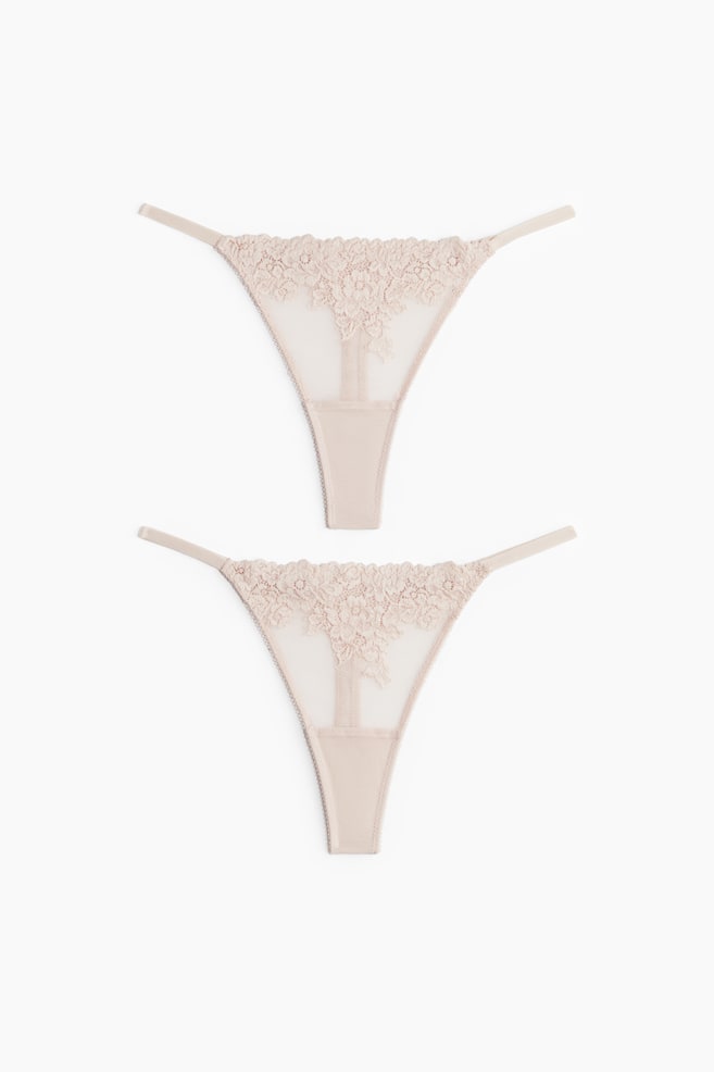 Dos tangas de color beige claro se muestran planas, mostrando su parte delantera. Cada tanga presenta un panel de encaje floral con bordes festoneados en la parte superior, que se transforma en un cuerpo de malla transparente con un ribete en zigzag a lo largo de los bordes, y un panel de entrepierna de tela sólida. Las finas tiras laterales se extienden desde la parte superior de encaje. - 1