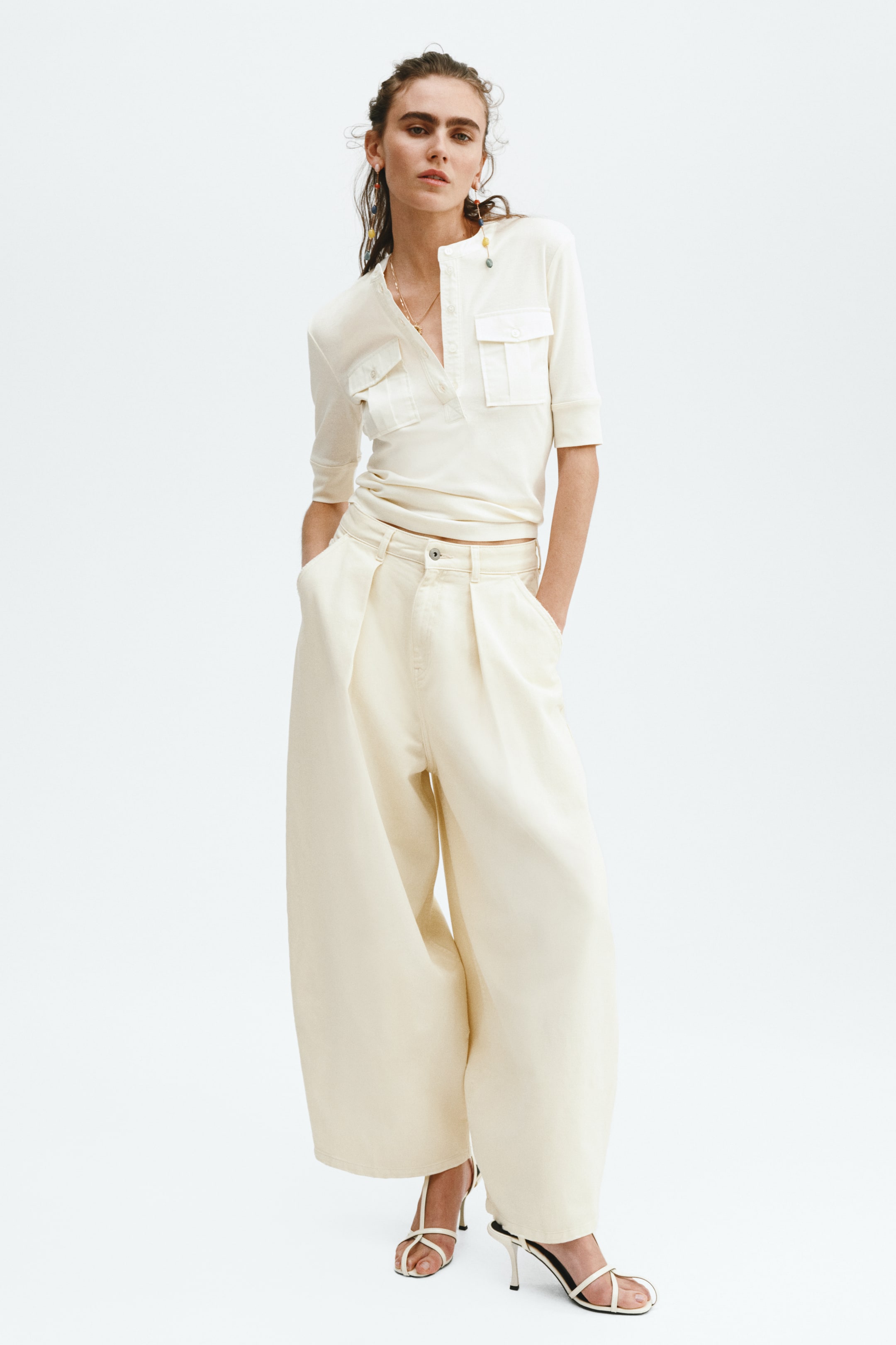 View larger image: A cream-coloured short-sleeved polo shirt, featuring two flap patch pockets and a V-neck placket, is being worn tucked into matching wide-leg trousers. The trousers are designed with front pleats and a single button fastening, and white strappy high-heeled sandals are completing the outfit, all presented facing slightly to the right.