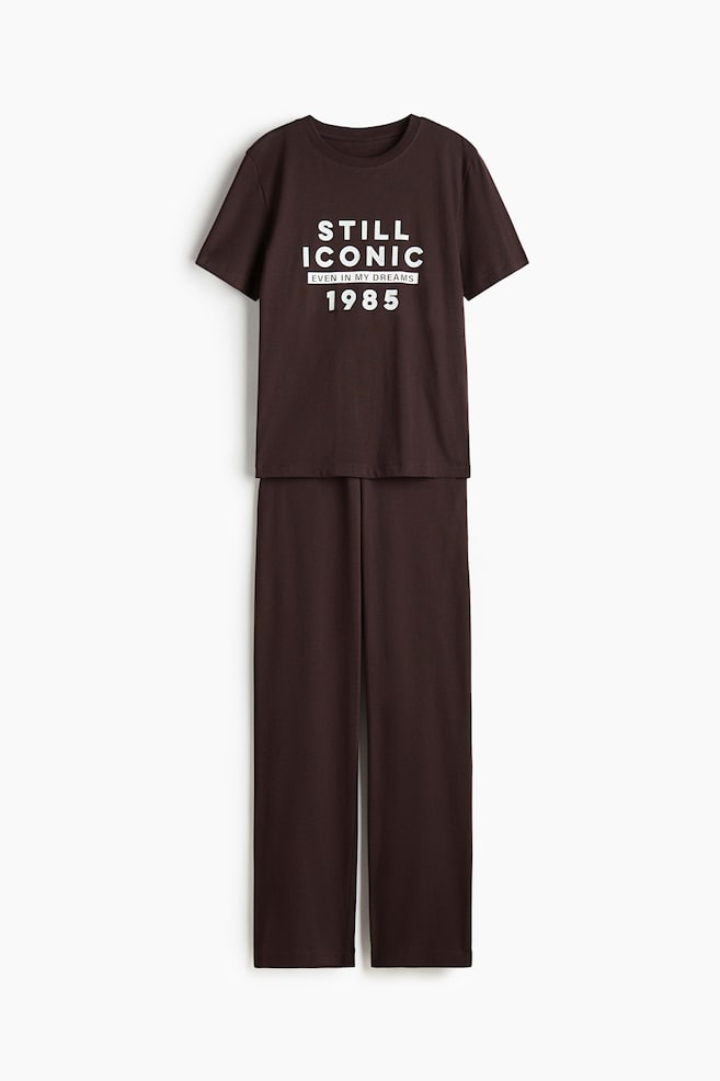 A set of brown jersey pyjamas is presented FRONT. The relaxed-fit top is featuring dropped shoulders, short sleeves, side slits, and a white "STILL ICONIC EVEN IN MY DREAMS 1985" graphic on its front. The matching bottoms are showing a covered elastic waist and straight hems. - 1