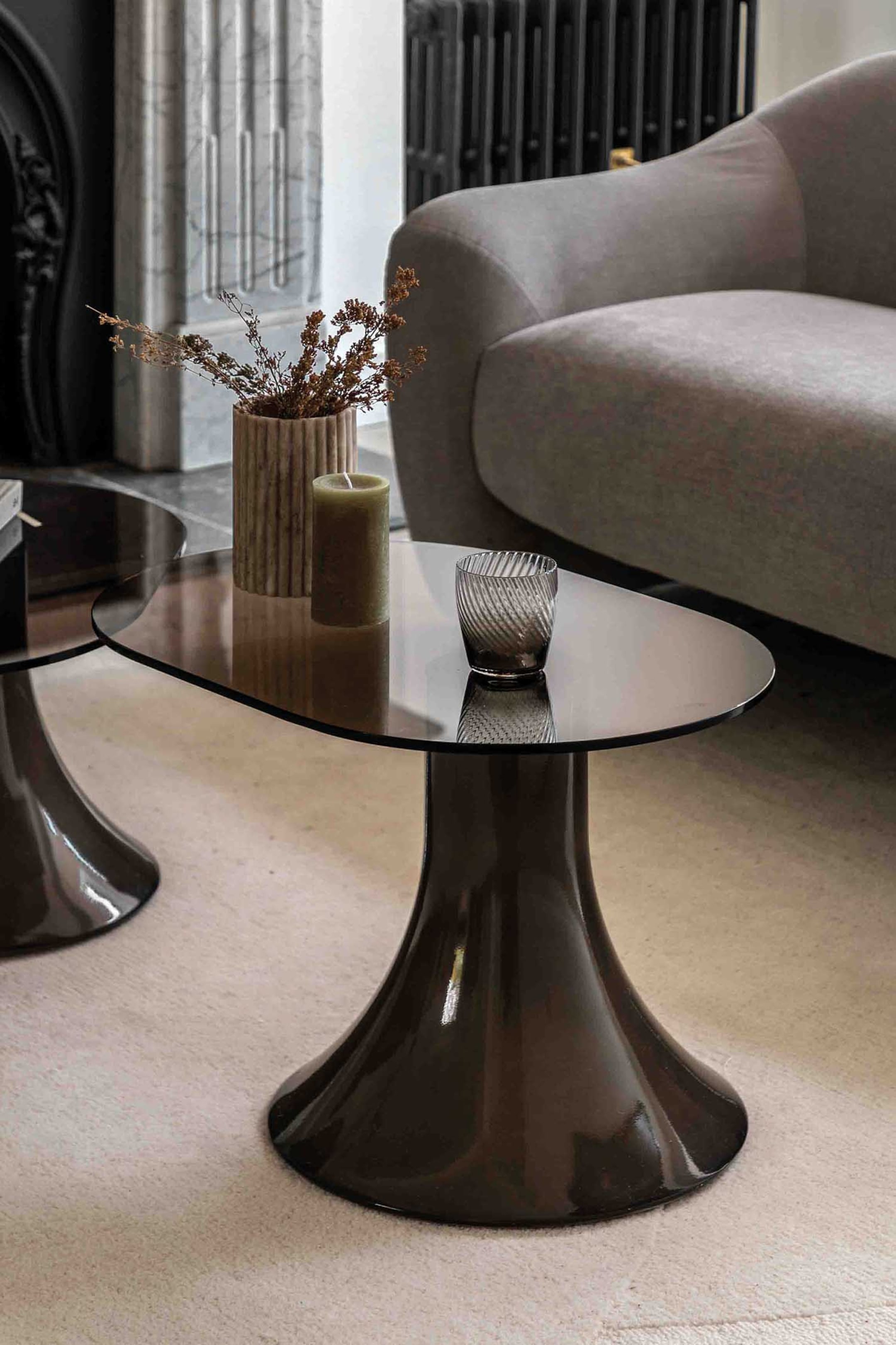 View larger image: Two coffee tables are showcasing irregular dark smoked glass tops and trumpet-shaped pedestal bases, with one table slightly overlapping the other. A ribbed vase holding dried botanicals, a green pillar candle, and a textured glass tumbler are resting on the larger table's surface.