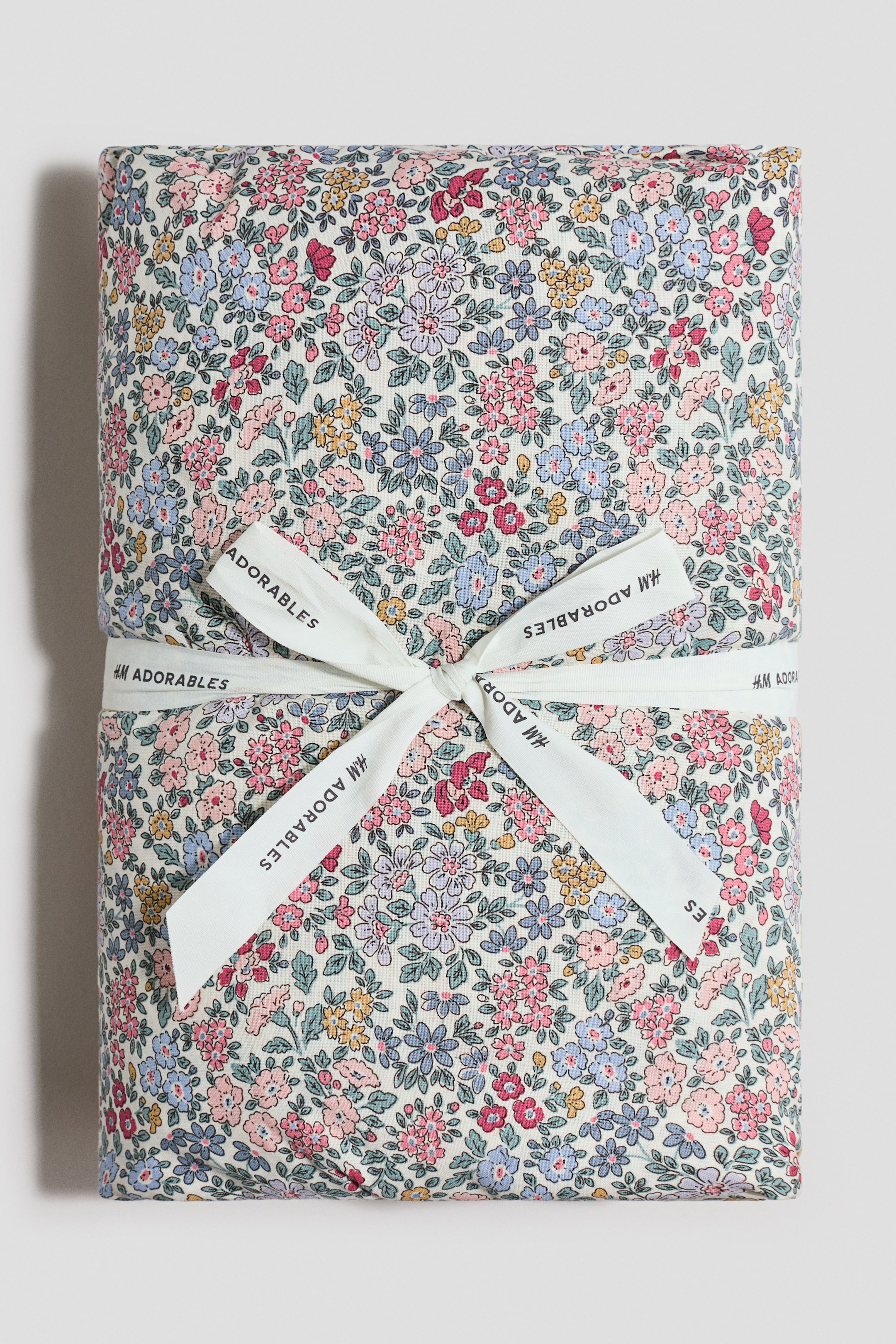 Agrandir l'image: Un article textile plié présente un motif floral dense avec des fleurs roses, bleues, jaunes et rouges sur un fond crème. Un ruban blanc, imprimé avec "H&M ADORABLES," est noué autour de l'article, formant un nœud à l'avant.
