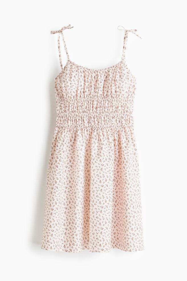 Se presenta un vestido corto desde el frente, confeccionado con una tela ligera y aireada con un pequeño estampado floral. Está diseñado con un escote profundo y redondo con elástico cubierto, tirantes estrechos con lazo en los hombros, fruncido en la cintura y una falda acampanada. - 1