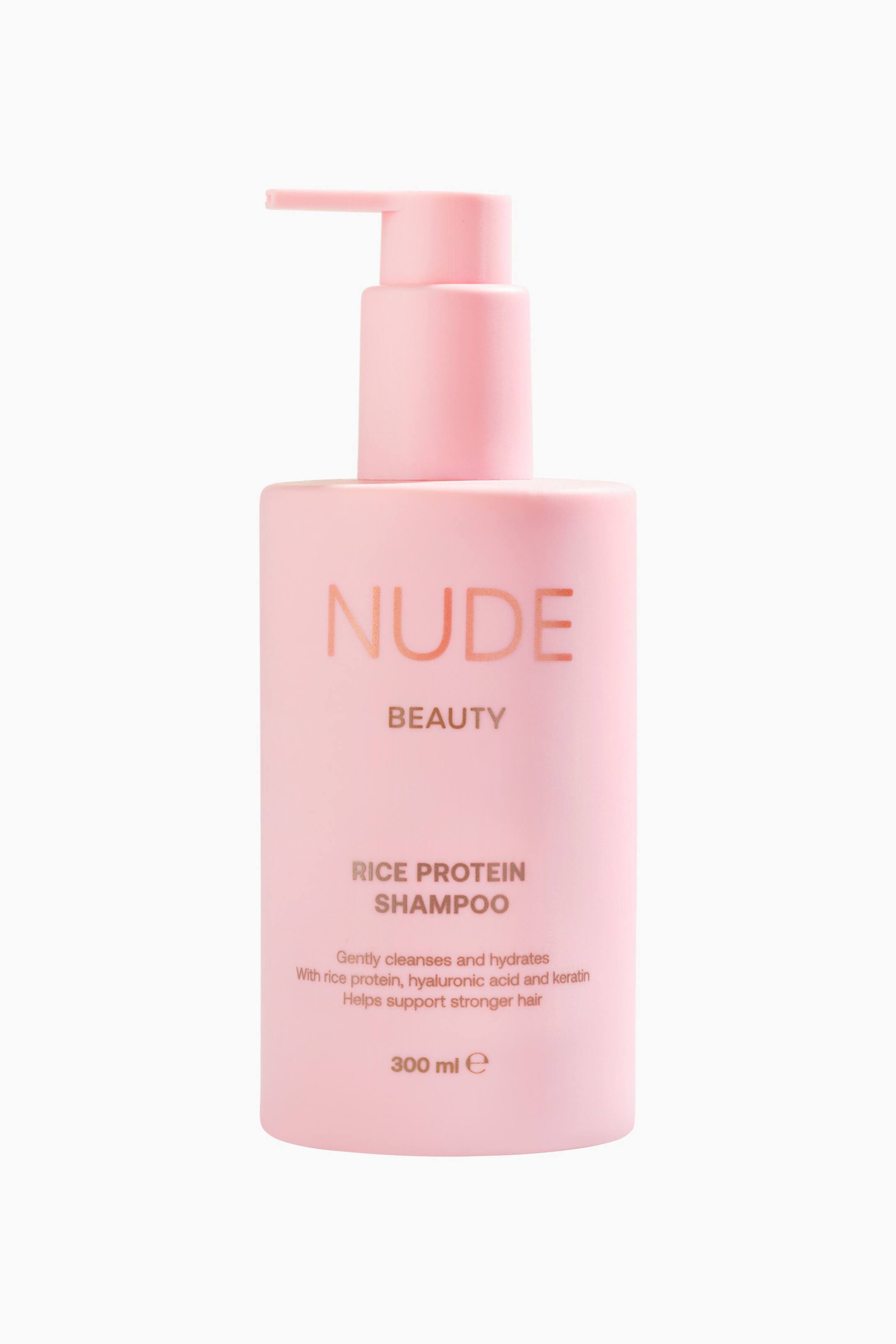 Vis større bilde: En rosa, rektangulær sjampoflaske med pumpe står oppreist og vendt fremover. Flasken viser "NUDE BEAUTY RICE PROTEIN SHAMPOO" i kobberfarget tekst, som beskriver fordelene og et volum på 300 ml.