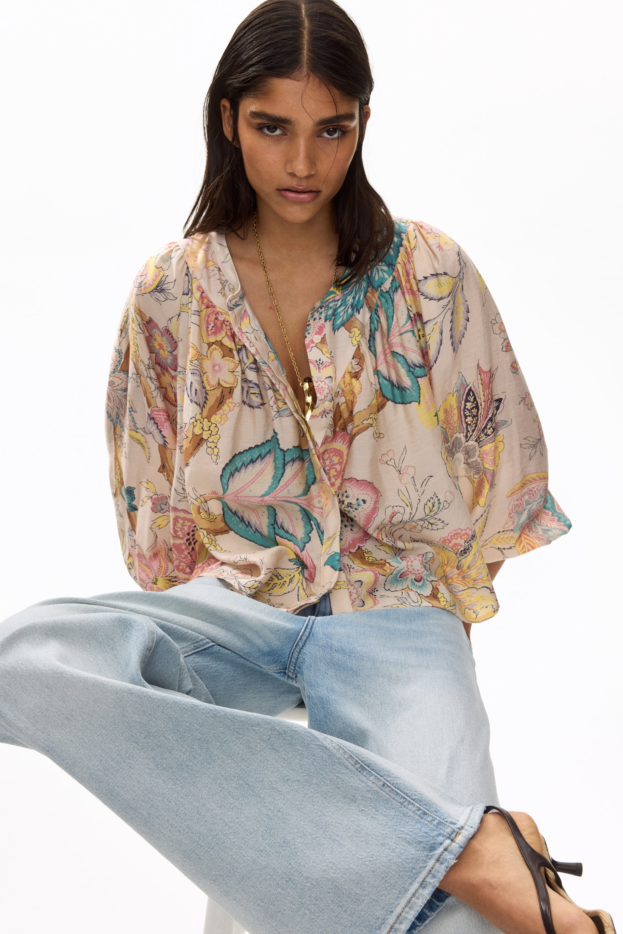 View larger image: The model is wearing a loose-fitting floral blouse adorned with pink, yellow, teal, and brown botanical designs. It features a band collar with a visible button, a front placket with concealed buttons forming a V-neck, a gathered V-shaped yoke, and long puff sleeves with buttoned cuffs. Complementing the blouse is a gold-toned necklace resting on the open neckline. Paired with the ensemble are light wash jeans, completing the look.