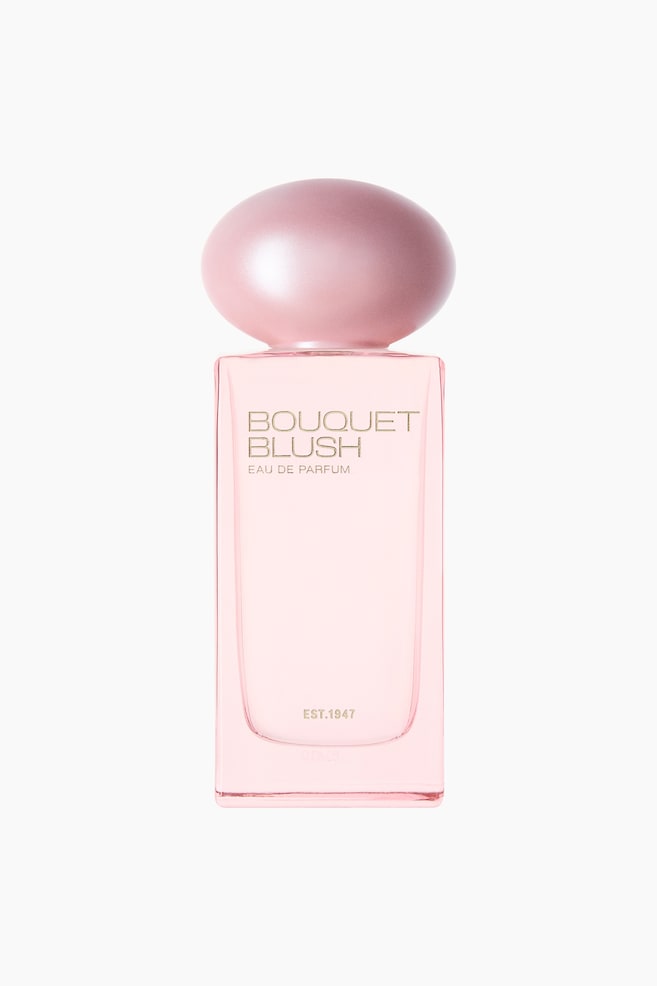 Bouquet Blush EdP prÃ¦senteres i en rektangulÃ¦r lyserÃ¸d glasflaske med en perlemorsskinnende, ovalformet lyserÃ¸d hÃ¦tte. Guldbogstaver er synlige pÃ¥ forsiden med teksten "BOUQUET BLUSH", "EAU DE PARFUM" og "EST. 1947". - 1