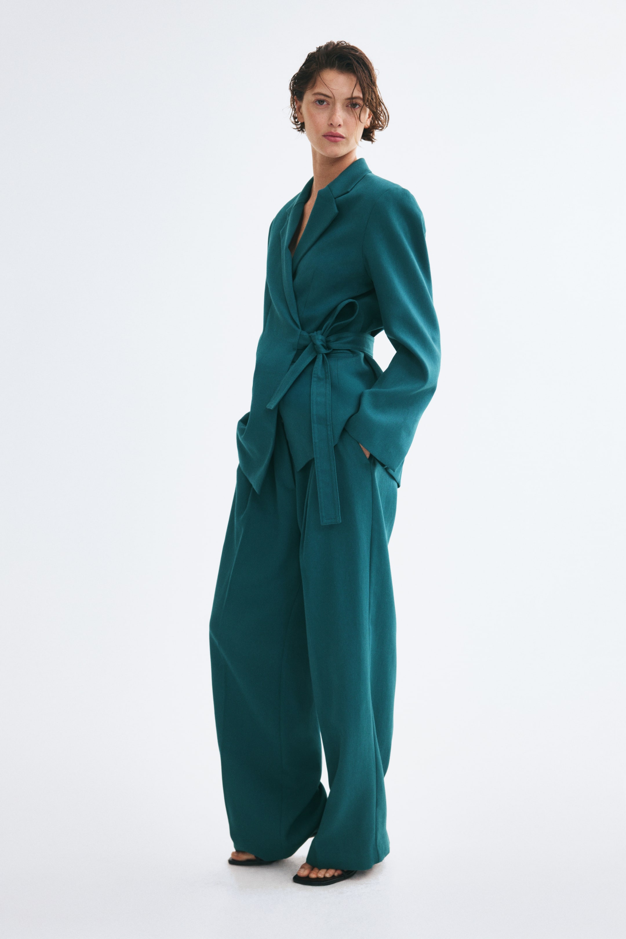 Agrandir l'image: Un pantalon ample vert bleu canard, taille haute et à jambes larges et droites, est porté par le mannequin avec un blazer portefeuille ceinturé assorti. Le mannequin est debout, la main glissée dans la poche latérale en biais.