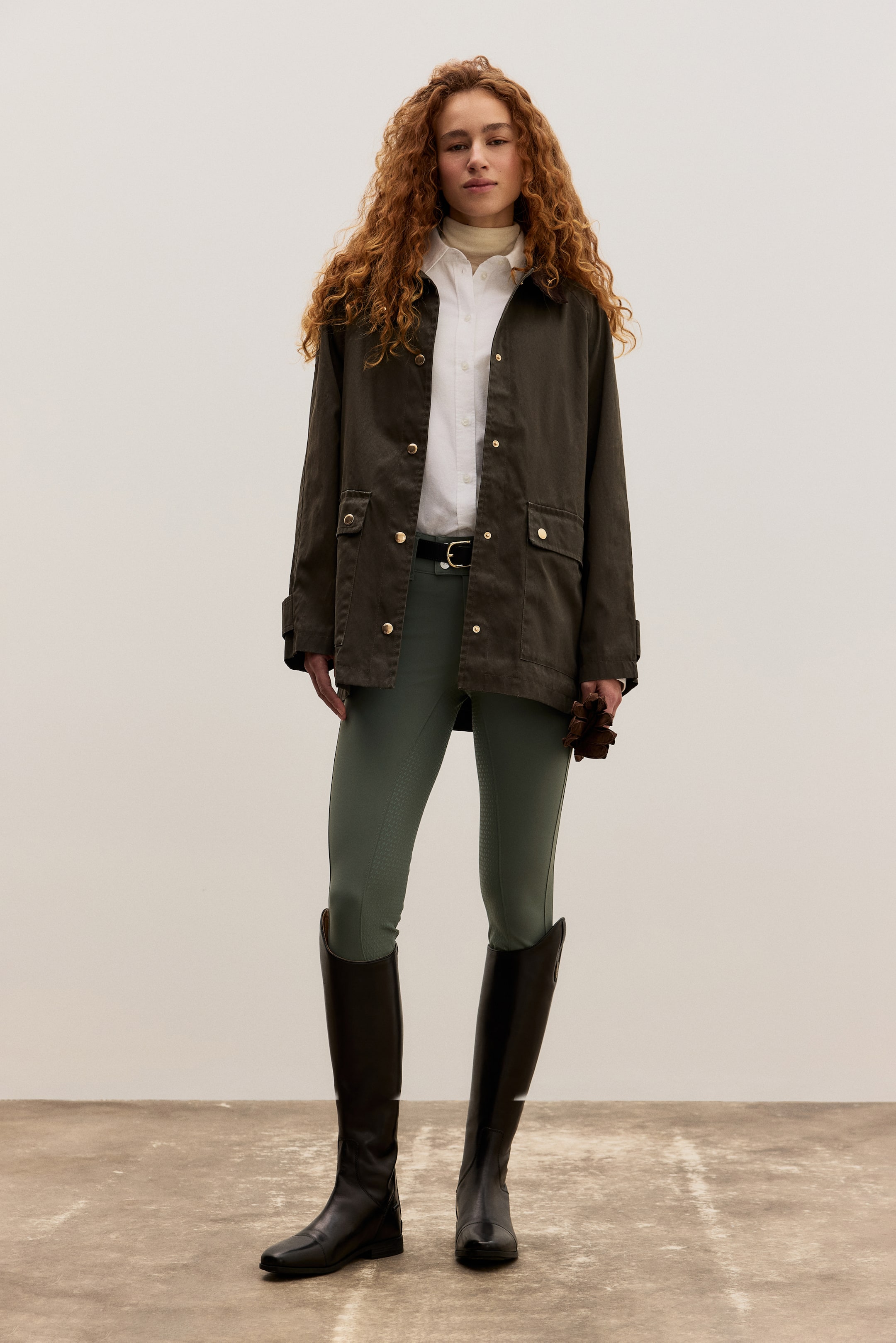 Grösseres Bild ansehen: Das Produkt ist ein Outfit, das nach vorne gerichtet präsentiert wird. Es besteht aus einer dunkeloliven Feldjacke mit goldfarbenen Druckknöpfen und Klappentaschen, die offen über einem weißen Hemd mit Kragen und einem cremefarbenen Rollkragenpullover getragen wird. In der Taille wird es mit einem schwarzen Gürtel zusammengehalten und vervollständigt den Look mit olivgrünen Reithosen, die strukturierte Griffflächen aufweisen, hohen schwarzen Reitstiefeln und braunen Lederhandschuhen, die in einer Hand gehalten werden.