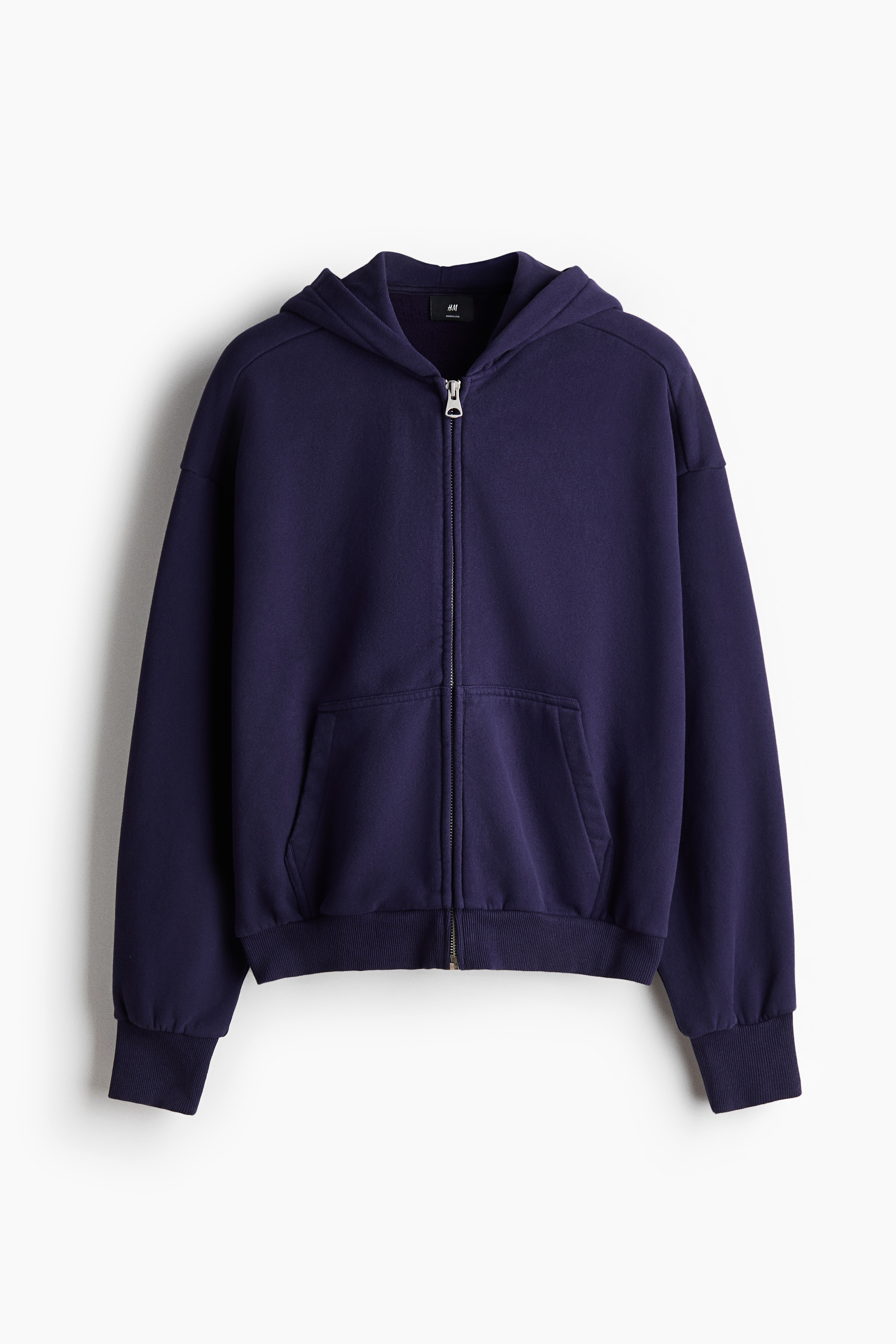 A dark purple zip-up hoodie in an oversized silhouette is worn by the model with their hands placed in the front pockets. The hoodie features dropped shoulders, a silver front zipper, and wide ribbed trim at the cuffs and hem, styled over a white crew-neck T-shirt.