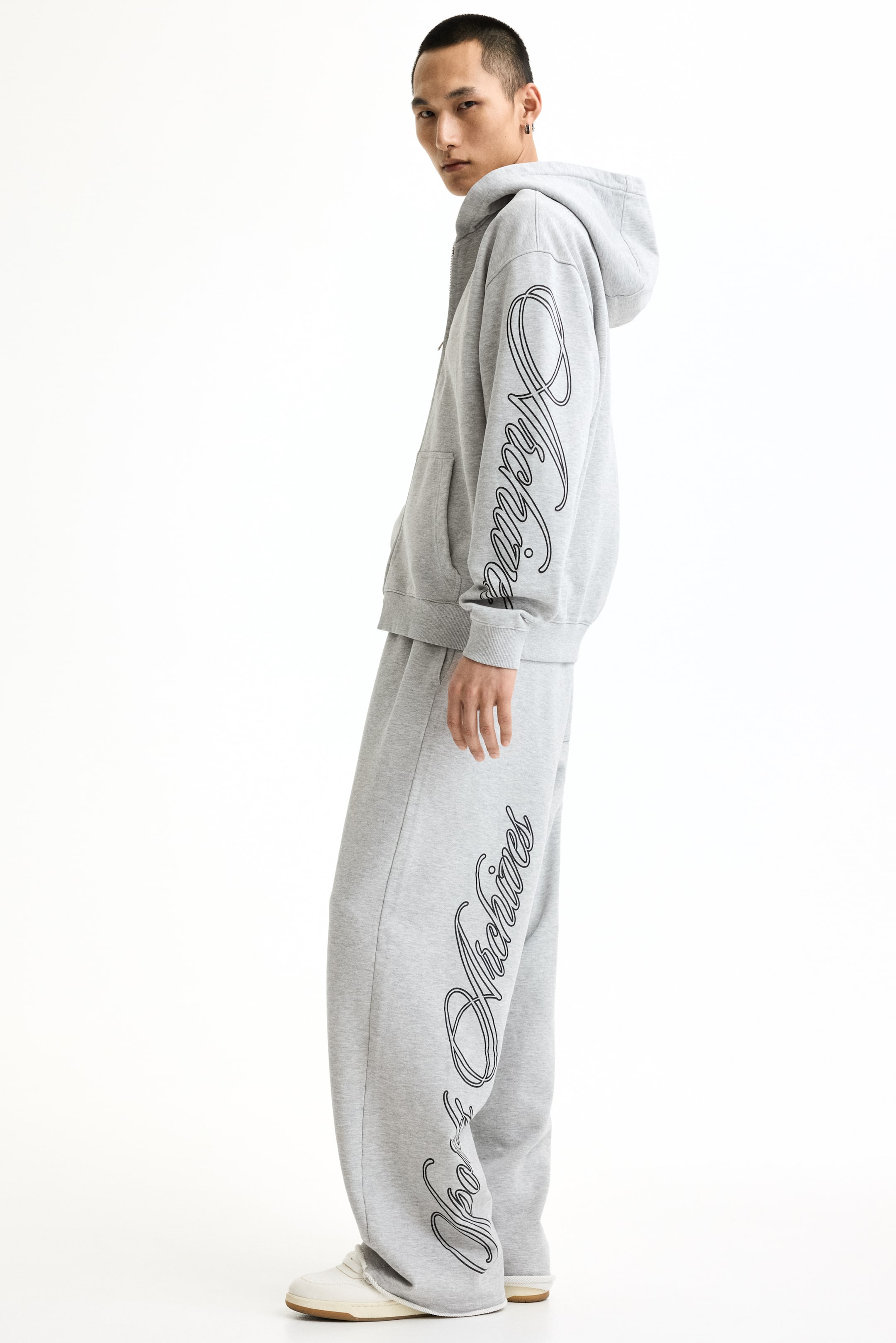 View larger image: A gray tracksuit is being showcased from a side angle, featuring an oversized hoodie with a kangaroo pocket and ribbed cuffs, paired with wide-leg sweatpants. Both the left sleeve of the hoodie and the left leg of the sweatpants display black script lettering, with the sweatpants also featuring a raw hem at the bottom.