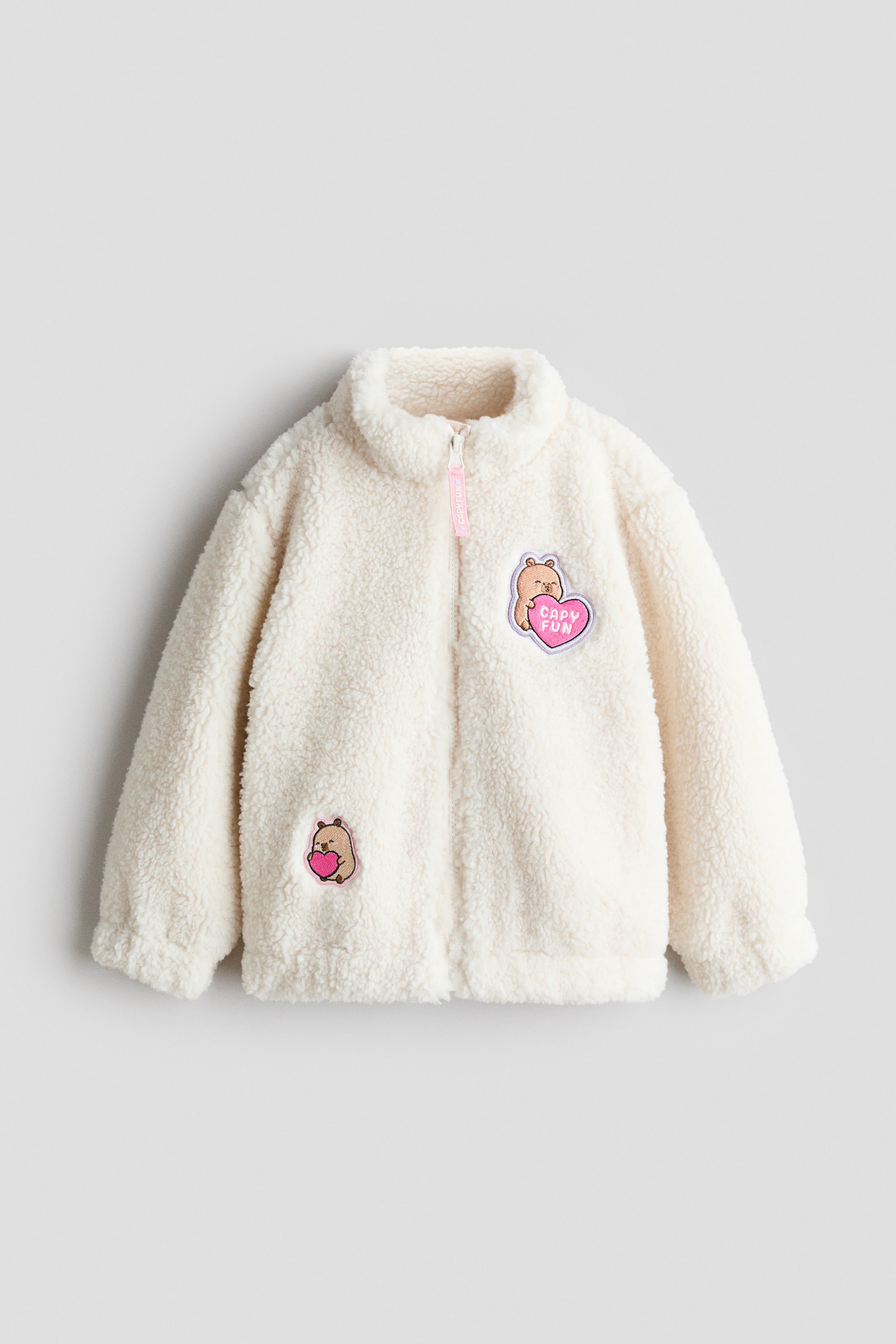 View larger image: A cream-colored teddy fleece jacket is shown from the front. It features a full-length zipper closure with a pink fabric tag, a stand-up collar, and long sleeves with elastic cuffs and hem. The jacket is accented with two embroidered patches: a larger patch on the upper right chest depicting a brown cartoon capybara next to a pink heart with the text "CAPY FUN," and a smaller patch on the lower left showing a capybara holding a pink heart.