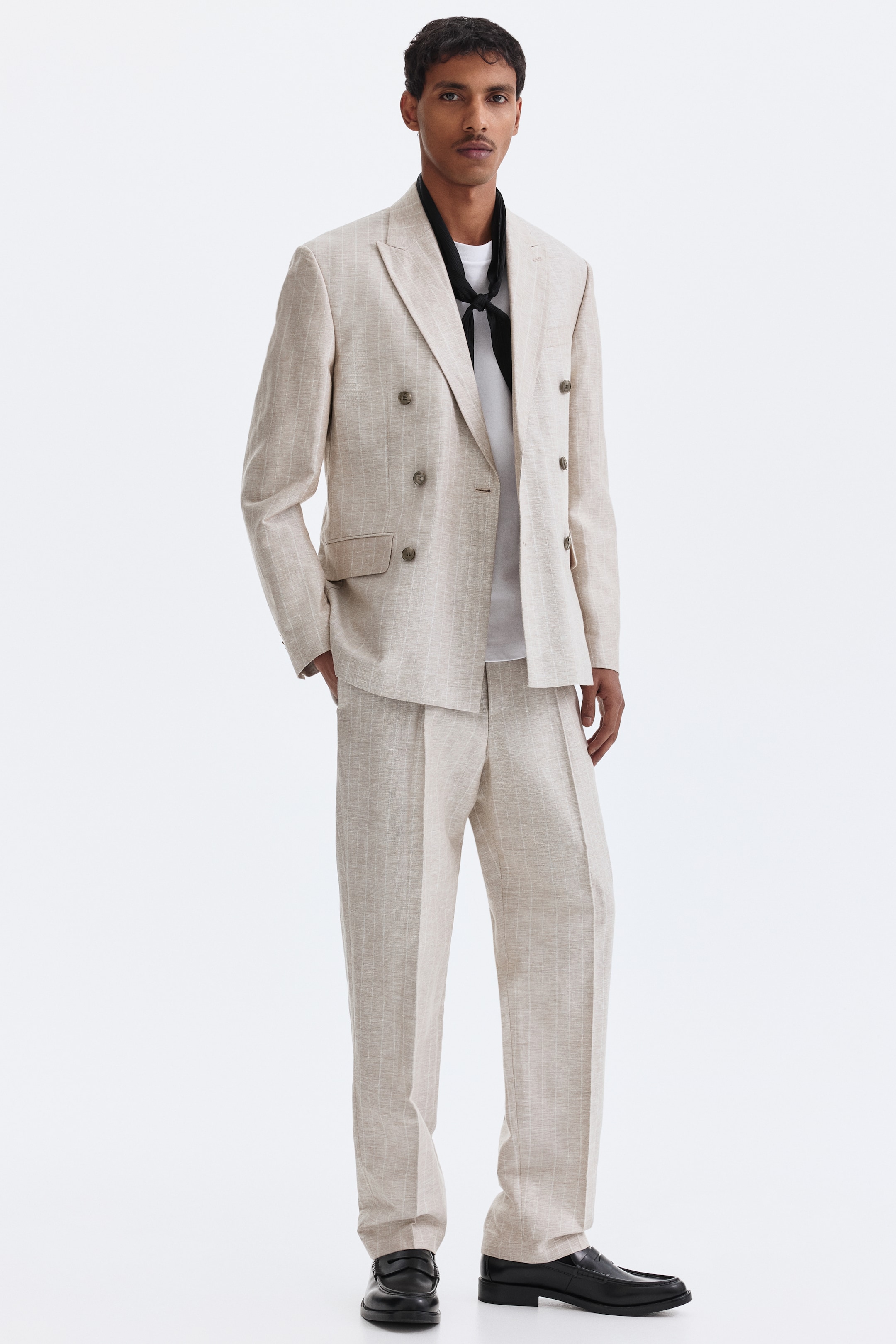 Nagyobb kép megtekintése: A modellen egy világos bézs, finom csíkos szövésű kétsoros zakó látható. Csúcsos hajtókákkal, hajtókás elülső zsebekkel és díszgombokkal a mandzsettán; fehér póló fölé viselve, hozzá illő nadrággal kombinálva. A modell az egyik kezét a zakó zsebébe helyezi.