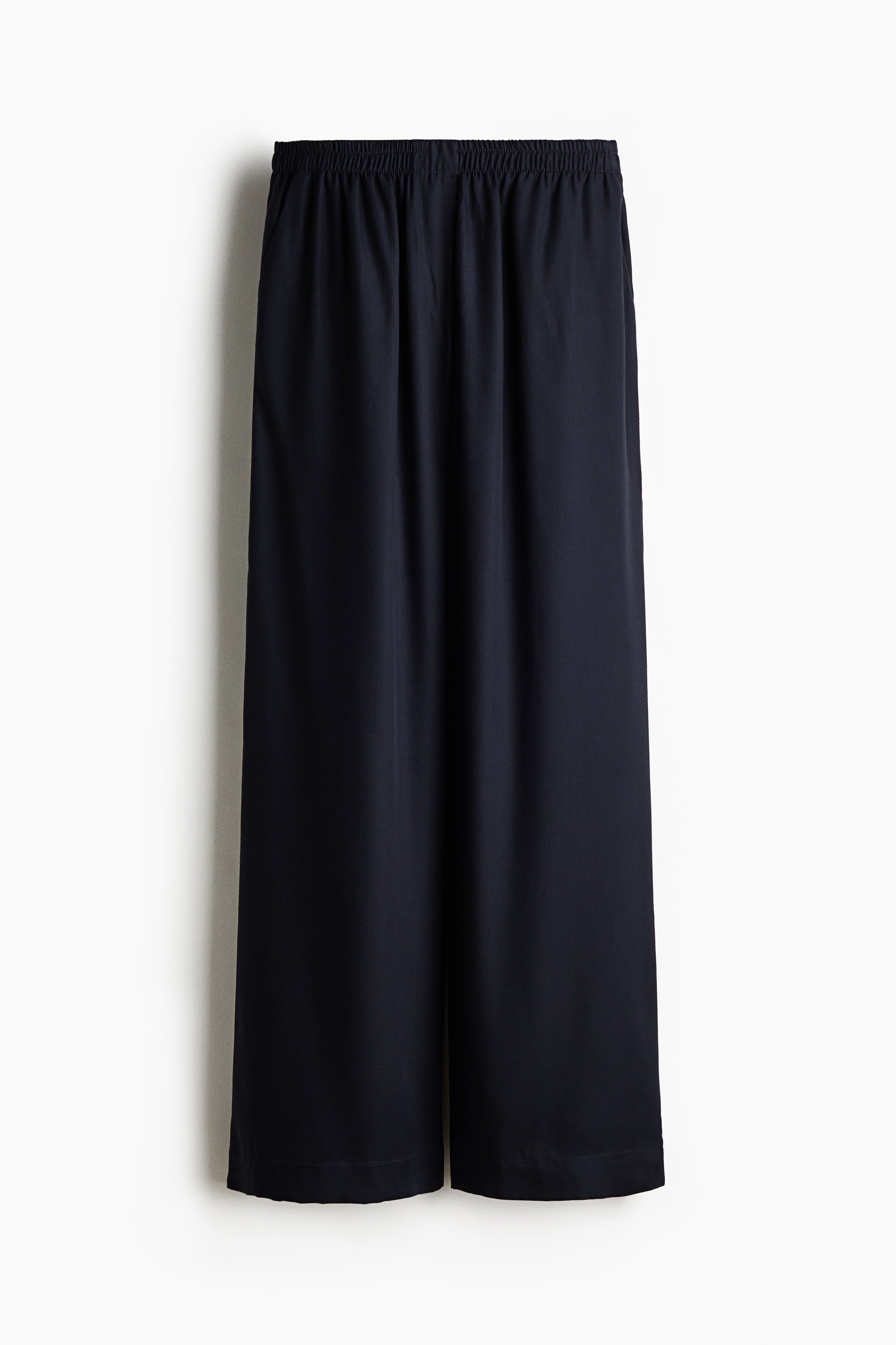 Die Hose aus dunklem Lyocell-Gewebe mit entspannter, weit geschnittener Silhouette und elastischem Bund wird vom Model getragen. Das Model steht mit den HÃ¤nden in den Seitentaschen und kombiniert die Hose mit einem passenden, aufgeknÃ¶pften Langarmhemd, das einen weiÃen Bund freilegt.