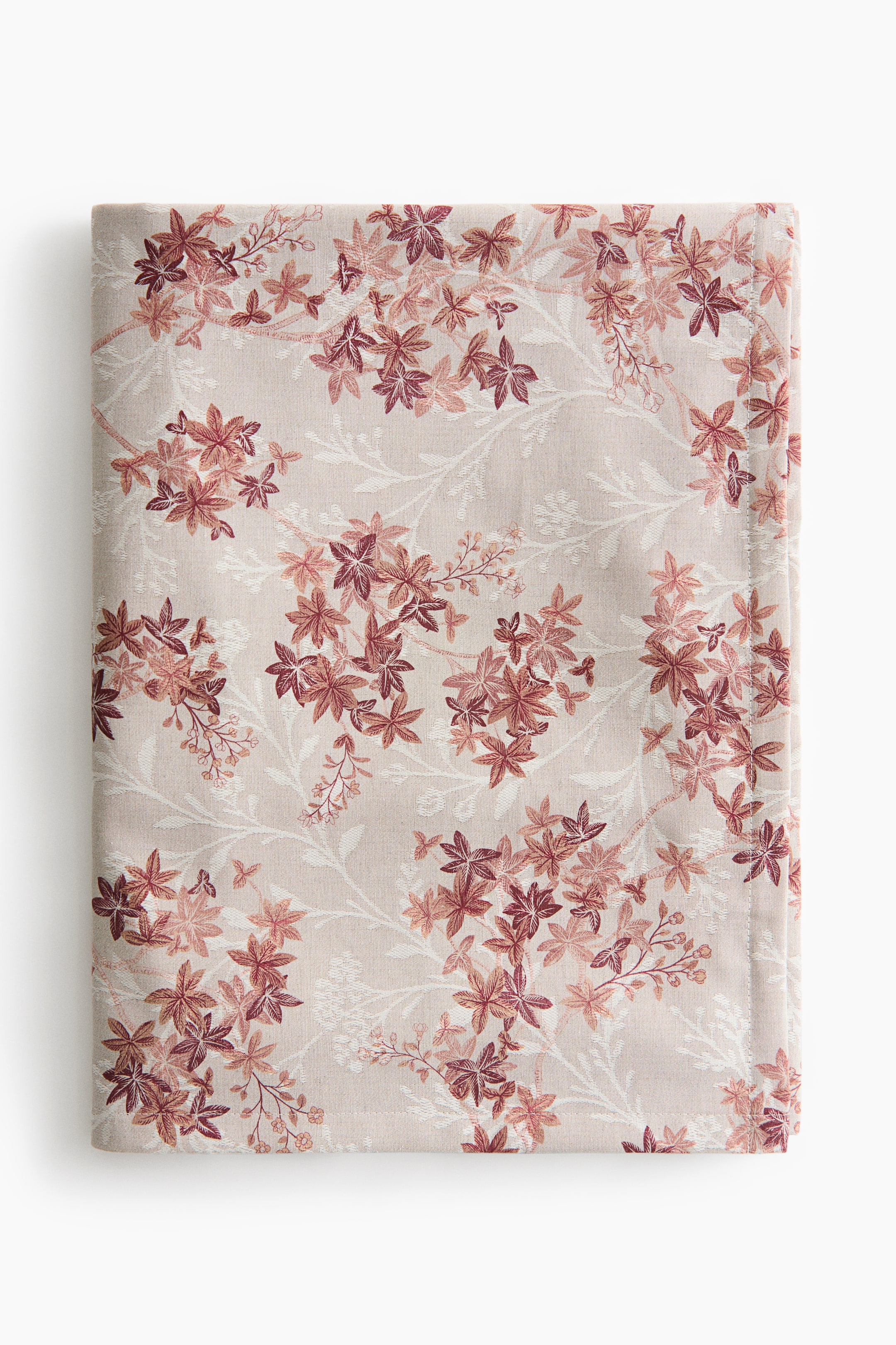 Grotere afbeelding bekijken: Een opgevouwen lichtbeige textiel toont een herhalend patroon van bladclusters in tinten poederroze, terracotta en bordeauxrood. De stof laat subtiele botanische silhouetten zien met witte contouren, afgewisseld tussen de gekleurde bladeren, met langs de rechterrand een nette gestikte zoom die zichtbaar is.