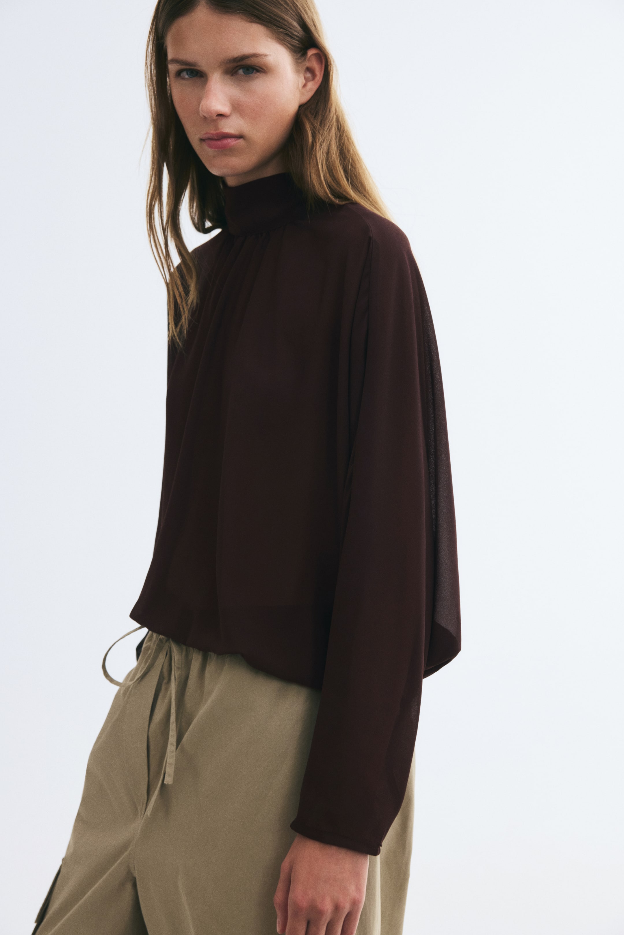 Ampliar la imagen: La modelo lleva una blusa marrón oscuro con cuello mao y mangas largas. La blusa ligera y con caída presenta un cuello alto fruncido y se combina con pantalones beige de cordón ajustable.