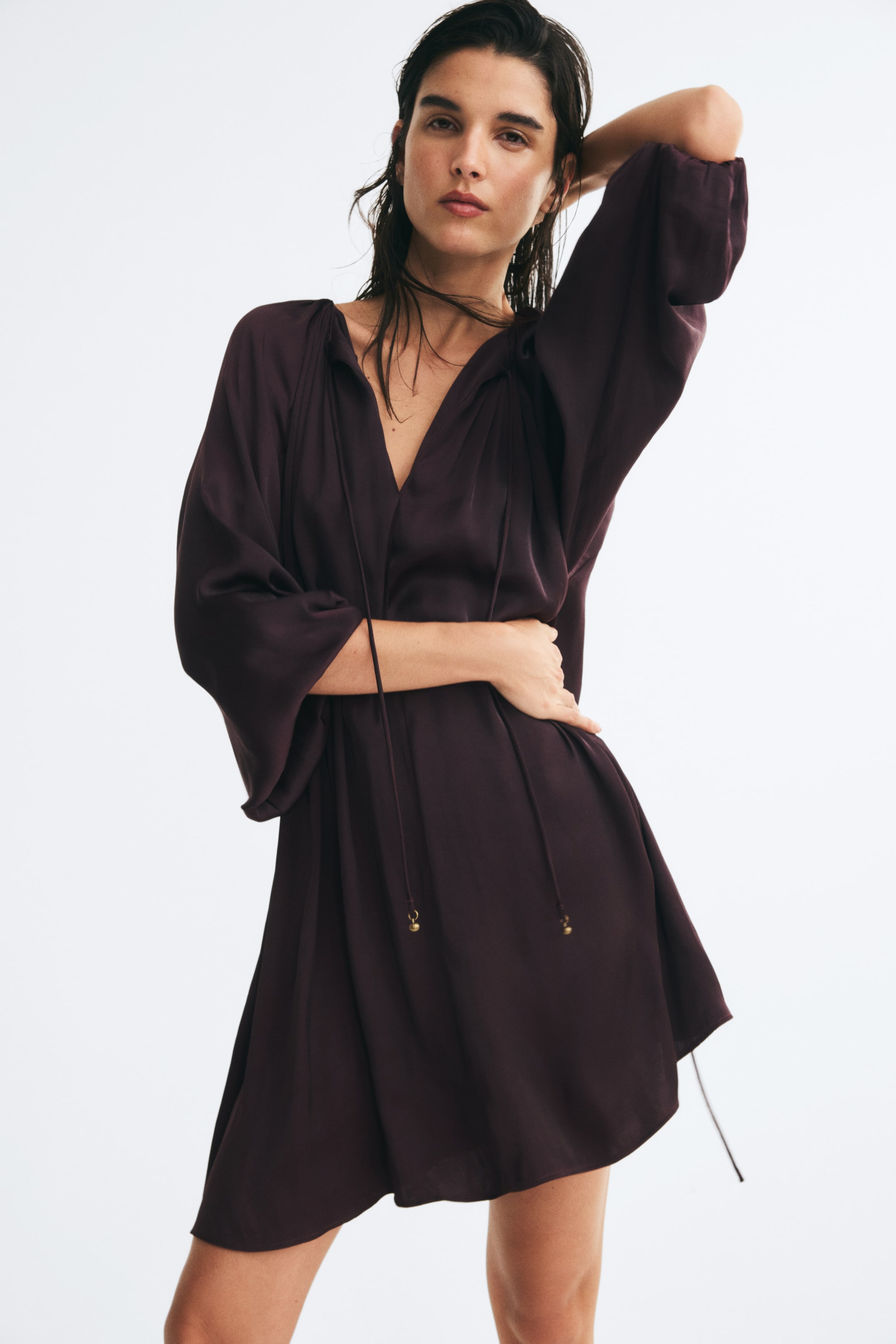 Voir une image plus grande: Une courte robe mauve foncé en tissu tissé légèrement brillant est portée par le mannequin, avec un décolleté en V muni de fines attaches perlées et de longues manches ballon drapées. Le mannequin pose avec une main sur la taille, mettant en valeur la taille froncée et la jupe évasée de la robe.