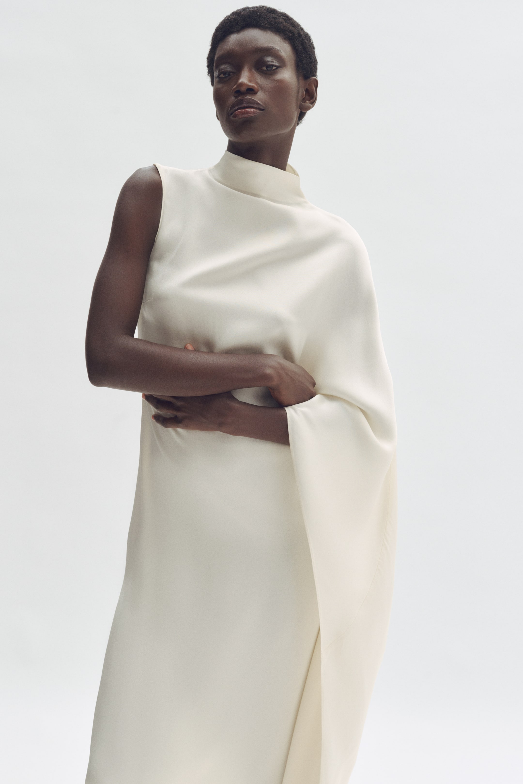 View larger image: An off-white dress featuring a high mock neck with an asymmetrical design. The left side presents a sleeveless cut, while the right side showcases a voluminous, flowing draped panel extending from the shoulder, creating a fluid silhouette.