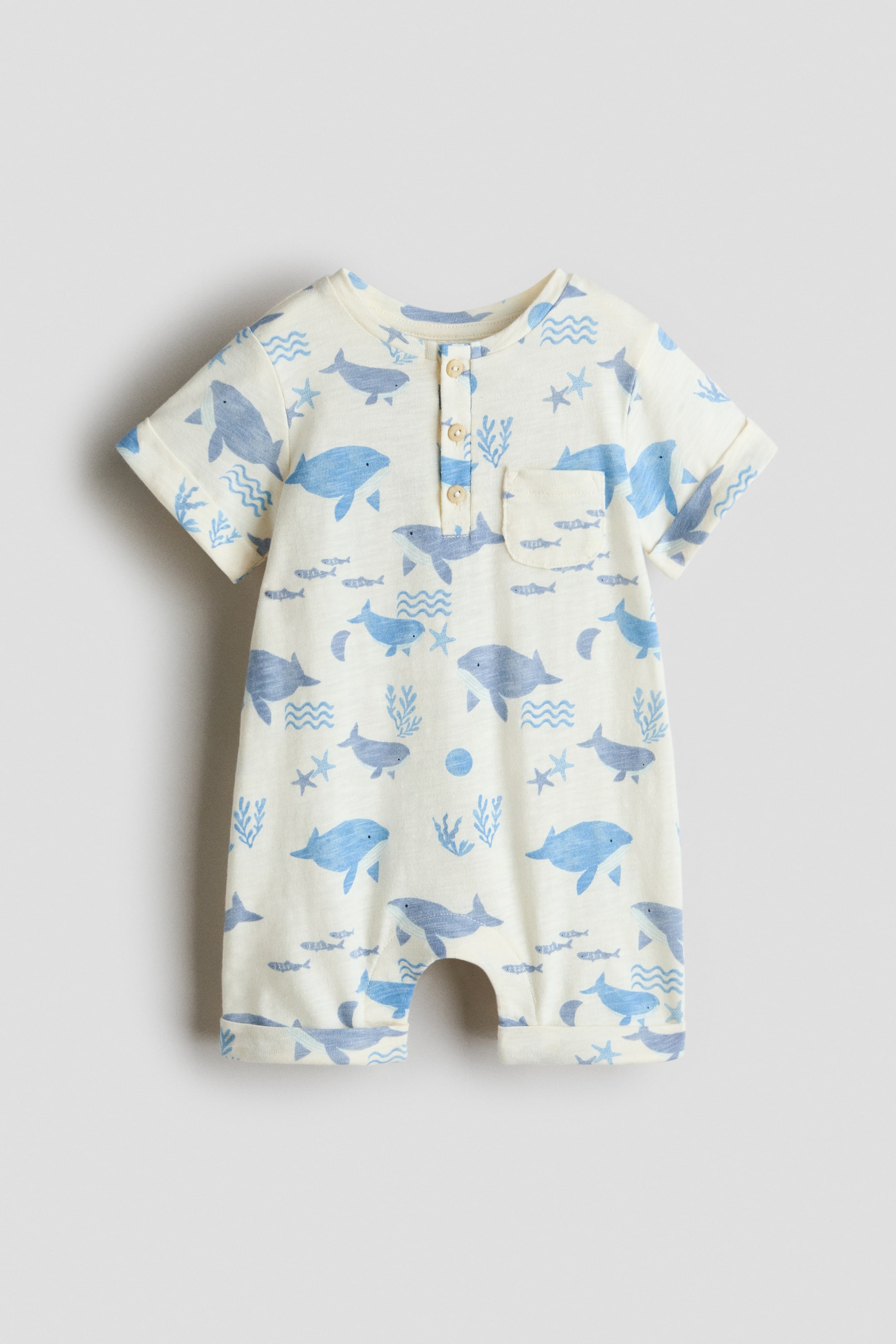 View larger image: The baby romper is shown from the front, crafted in a cream fabric with an all-over blue whale and sea-themed print. It features short, rolled sleeves, a three-button placket at the round neckline, a single chest patch pocket and short legs with rolled cuffs.