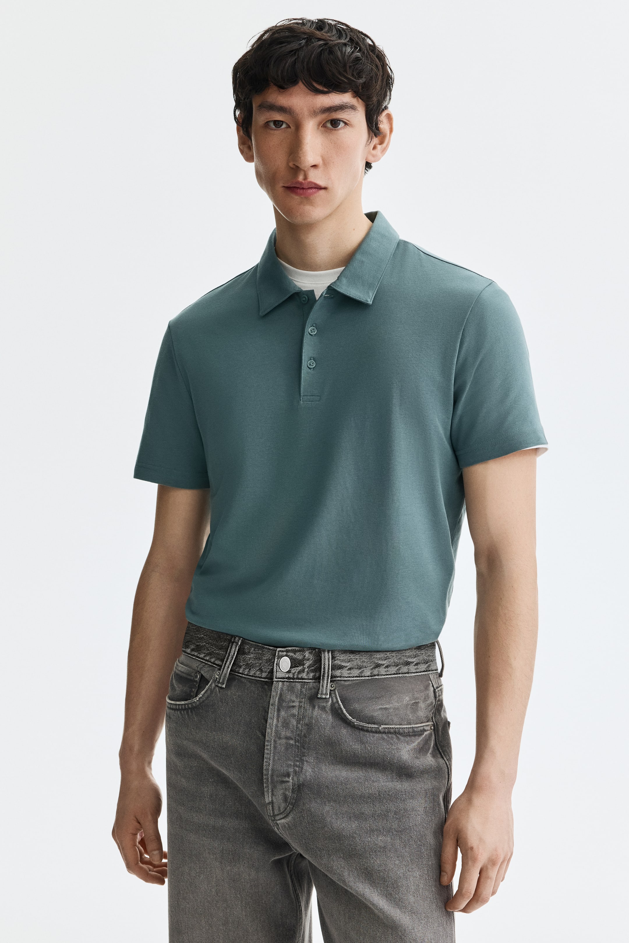 View larger image: A dark teal short-sleeved polo shirt is being worn front-facing, featuring a flat collar and a three-button placket with dark buttons. A white t-shirt is layering underneath, visible at the collar and sleeve hems.