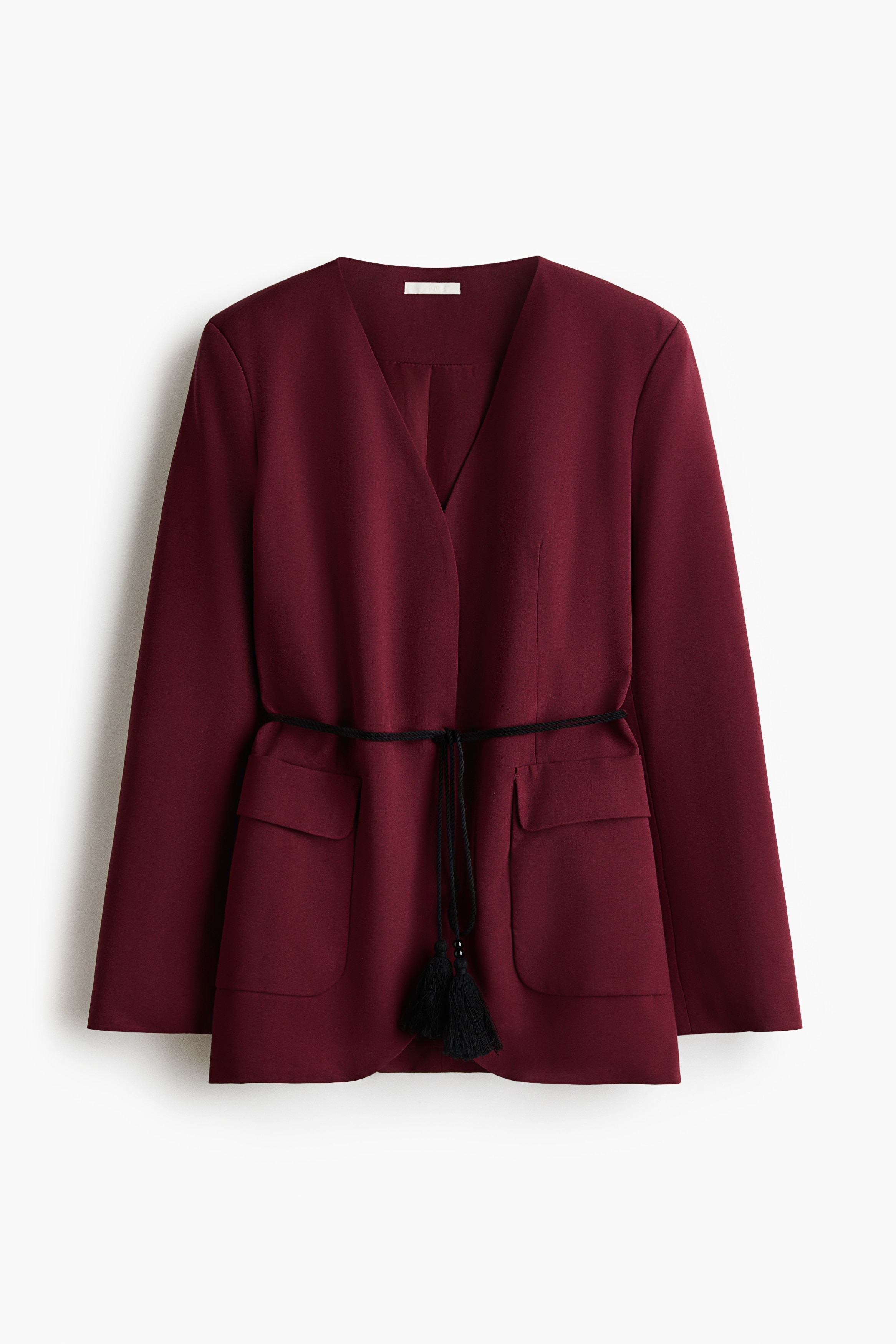 This burgundy, lapelless blazer is presented from a three-quarter front angle, featuring structured shoulder pads, a deep V-neckline, and front flap pockets. The blazer is styled closed with a thin black twisted rope tie belt with tasseled ends at the waist, worn over wide-leg light blue jeans.