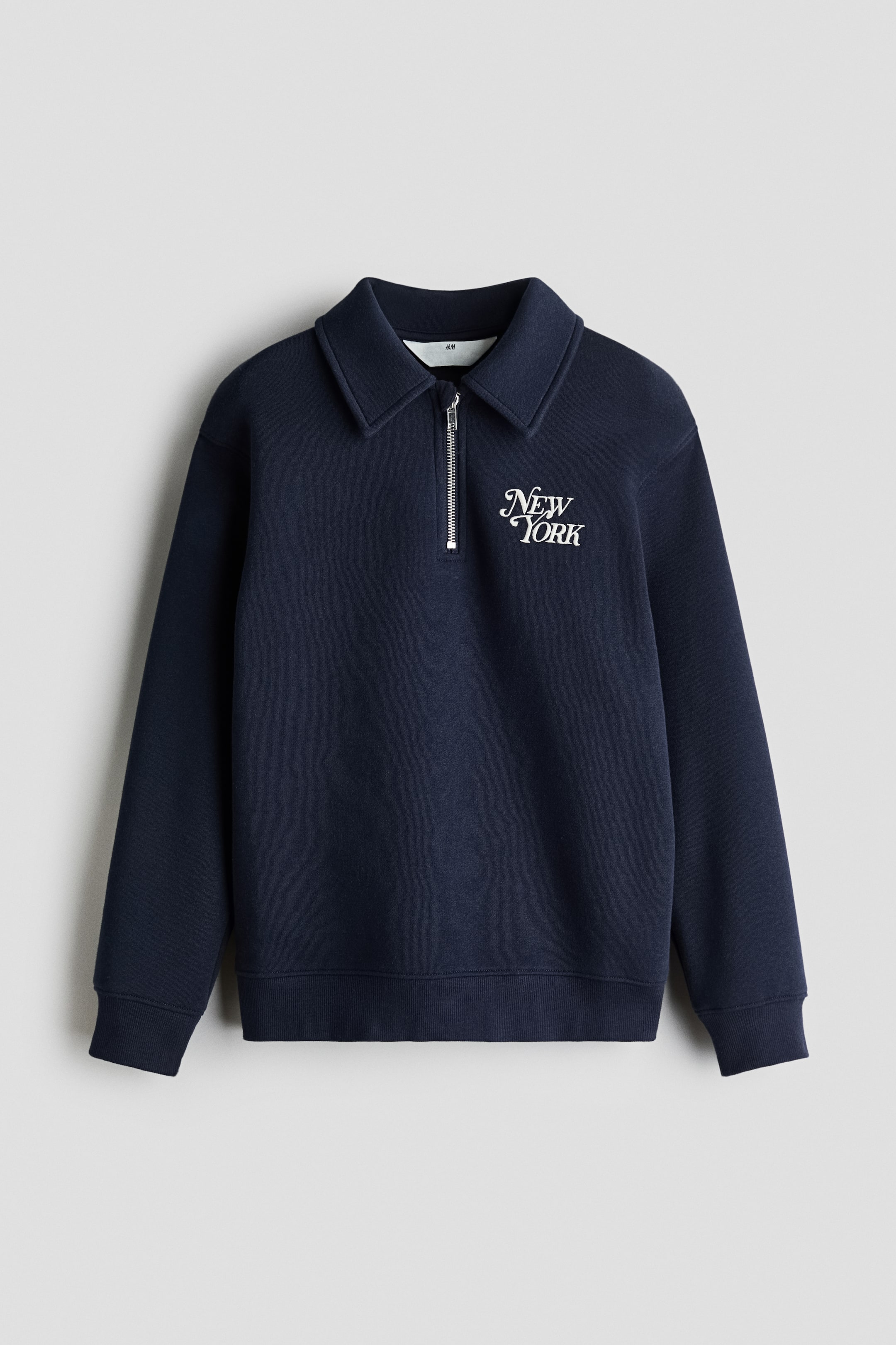 View larger image: A navy blue half-zip sweatshirt is shown front-facing, featuring a structured polo collar, a silver-toned metal zipper, and the words "New York" embroidered in white script on the left chest. The garment features long sleeves with ribbed cuffs and a matching ribbed hem.