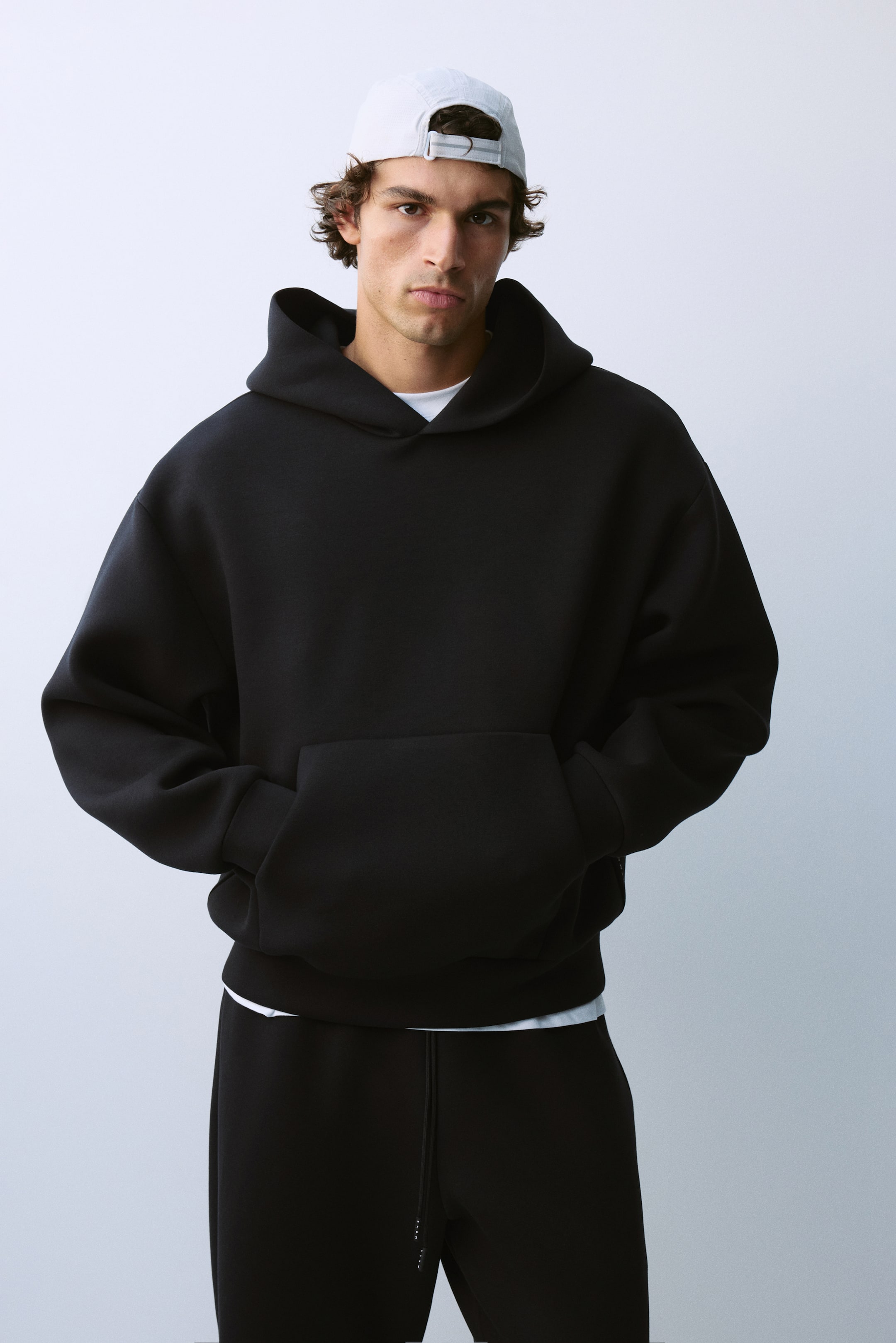 Zobrazit větší obrázek: Model má na sobě černou oversized mikinu s kapucí se spuštěnými rameny a přední klokaní kapsou, přičemž obě ruce má položené v kapse. Mikina je vrstvená přes bílé tričko a doplněná ladícími černými tepláky.