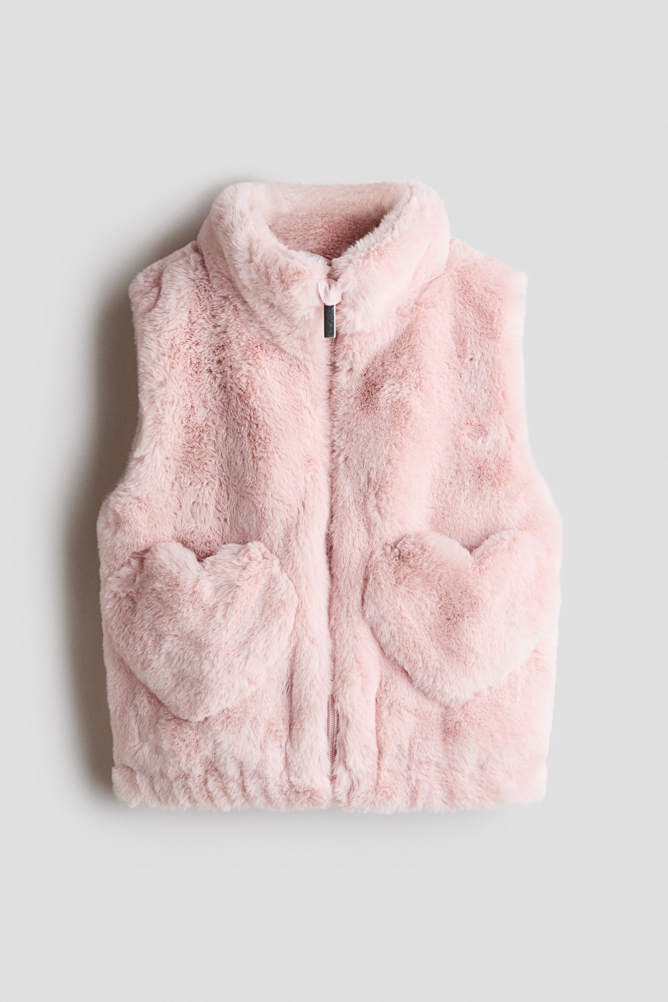 Visualizza immagine più grande: Un gilet in ecopelliccia rosa chiaro è ripreso frontalmente, mettendo in mostra un colletto rialzato e una chiusura frontale completamente zip con un piccolo proteggi-mento. Il gilet presenta due tasche applicate a forma di cuore nella parte inferiore anteriore e un orlo elasticizzato.