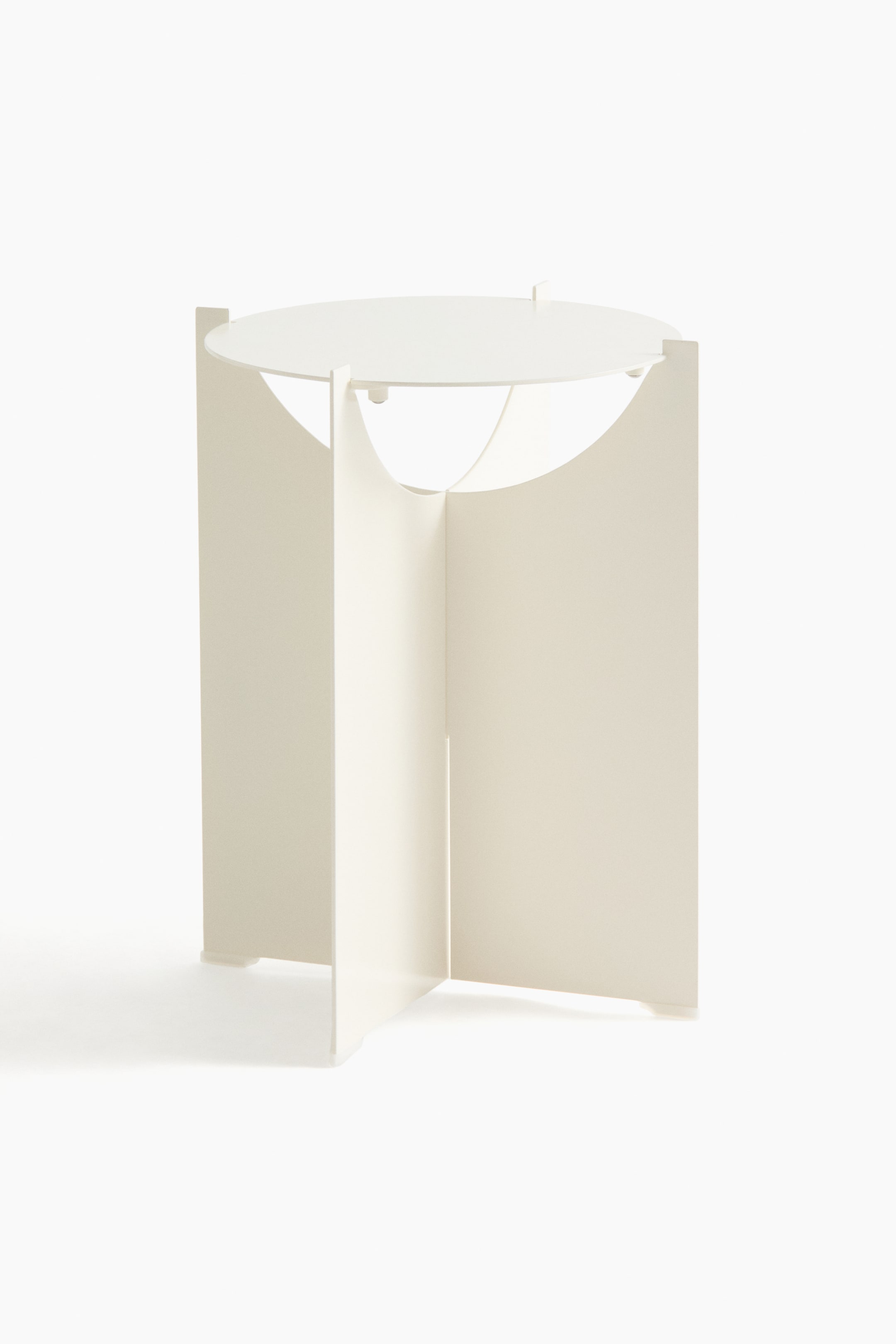 View larger image: A side table, front view, highlights a minimalist design featuring a round, cream-coloured stainless steel tabletop. The tabletop is supported by an interlocking base formed by four flat panels, each with a curved upper edge.
