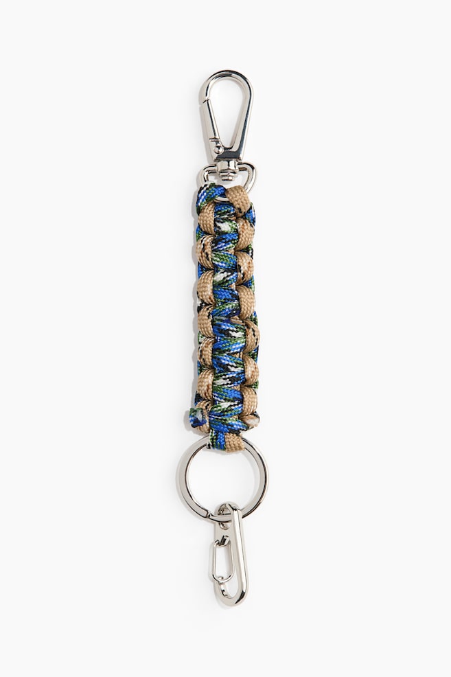 Un charm pour sac présente un cordon tressé dans des couleurs bleu, vert, noir, blanc et beige, disposé verticalement. Un mousqueton argenté apparaît en haut, et un anneau porte-clés avec un petit mousqueton supplémentaire est visible en bas. - 1