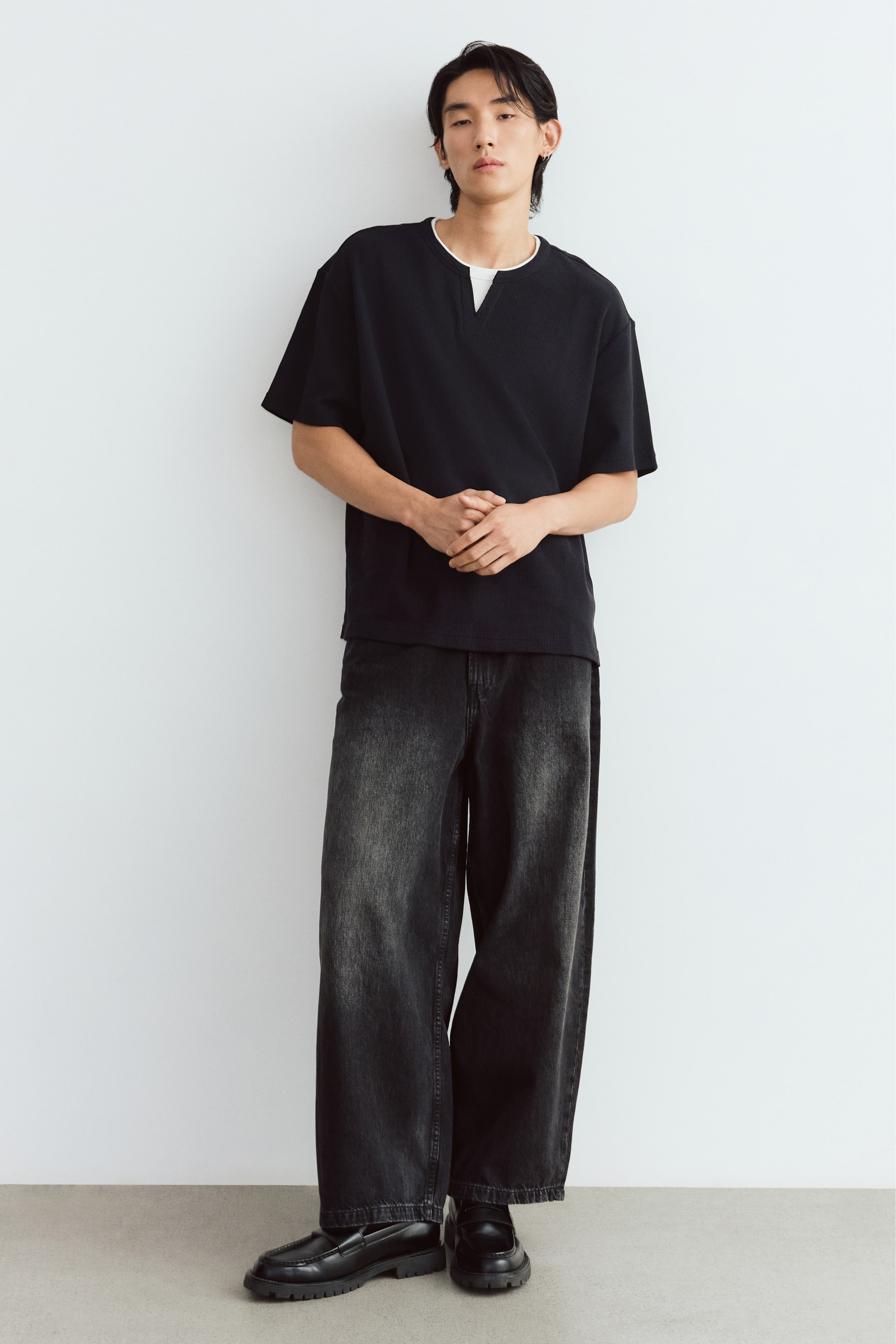View larger image: The model is wearing a black waffle-knit jersey tee with a layered look, featuring a visible white collar, a split round neckline, and dropped shoulders. The loose-fit tee is styled with dark-wash wide-leg denim jeans and black leather loafers.