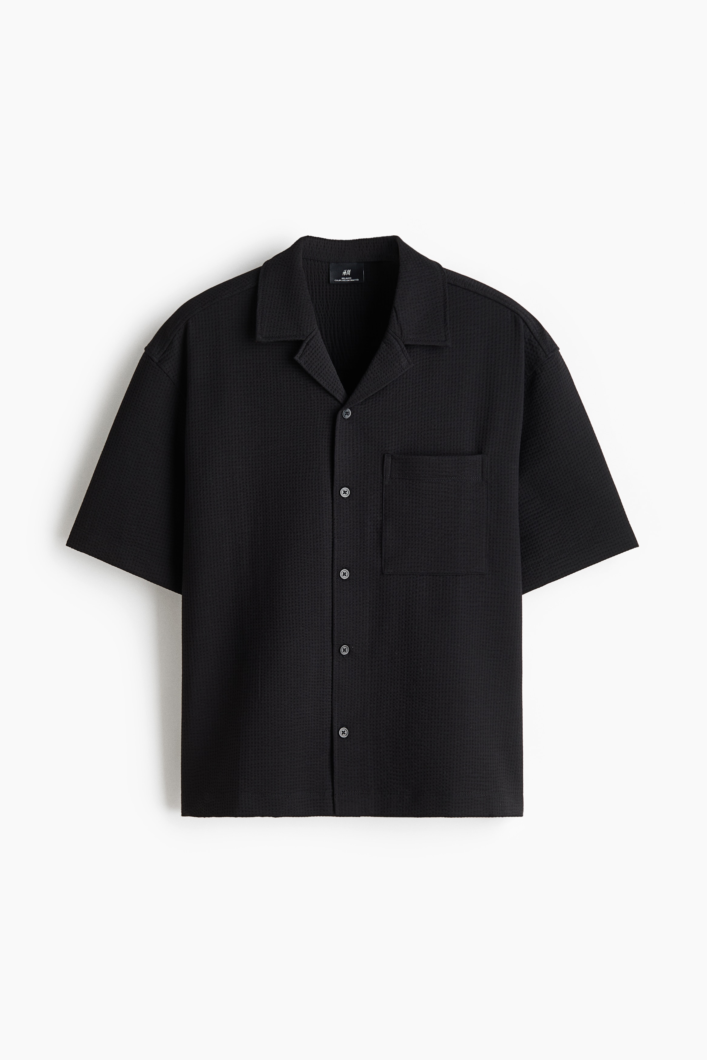 A black, short-sleeved shirt in a waffled cotton blend is worn open by the model, showcasing its relaxed fit, resort collar, and single chest pocket. The shirt is styled over a black tank top and dark trousers with a silver-buckled belt.
