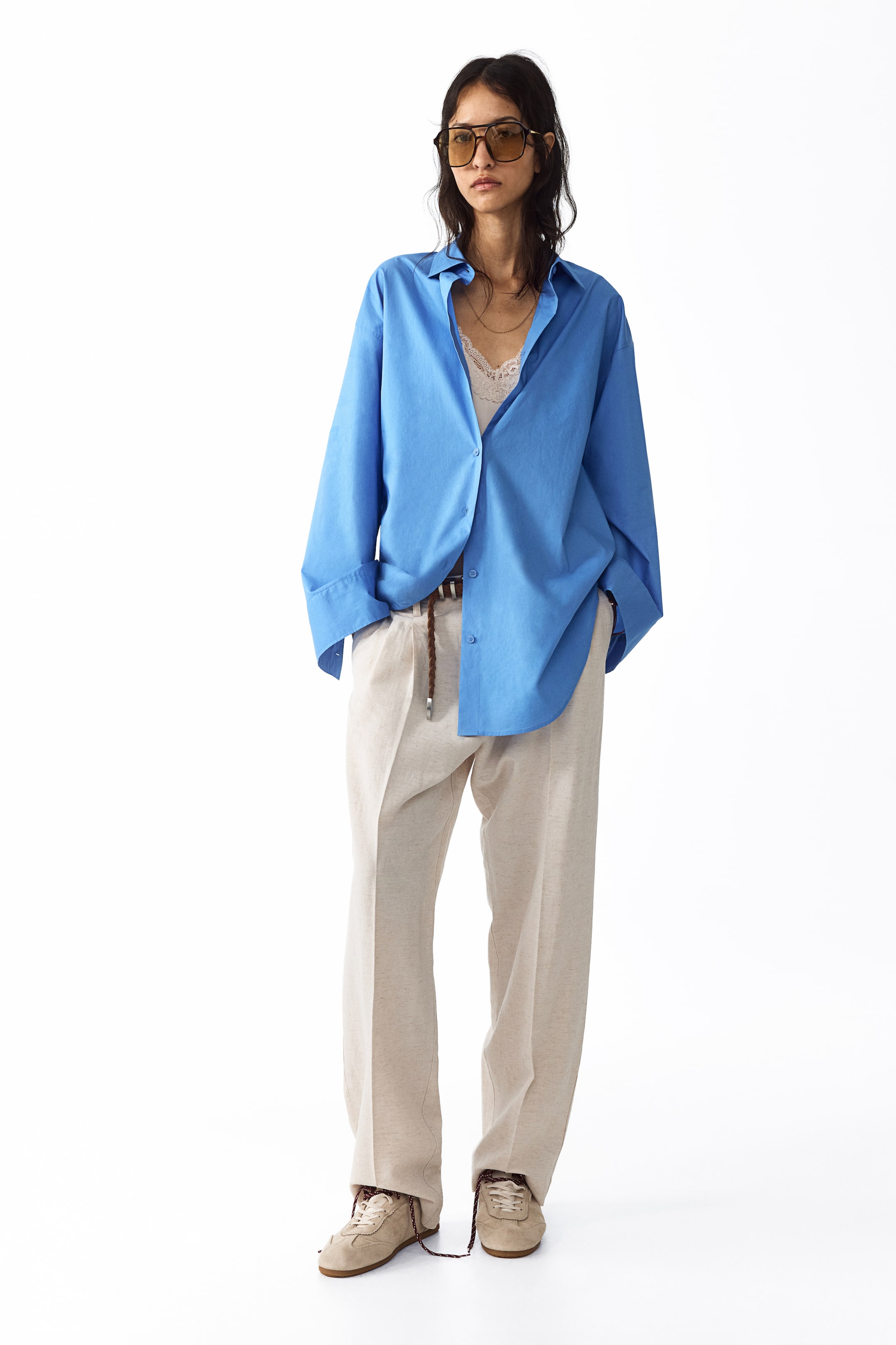 View larger image: The model is wearing light-colored, loose-fit trousers made from a linen-blend weave. These trousers showcase front pleats and visible creases, with the model's hands comfortably placed in the diagonal side pockets. The waist is elegantly cinched with a braided belt.