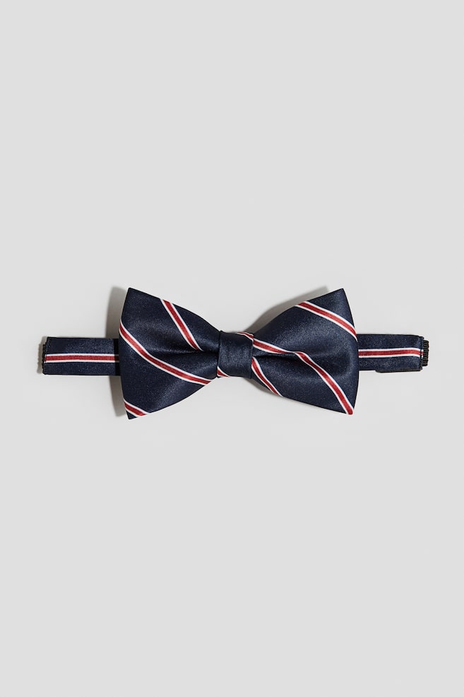 A pre-tied bow tie is being shown from the FRONT, featuring a navy blue woven fabric with diagonal red and white stripes. An adjustable elastic strap with a black plastic fastener is extending from its sides. - 1