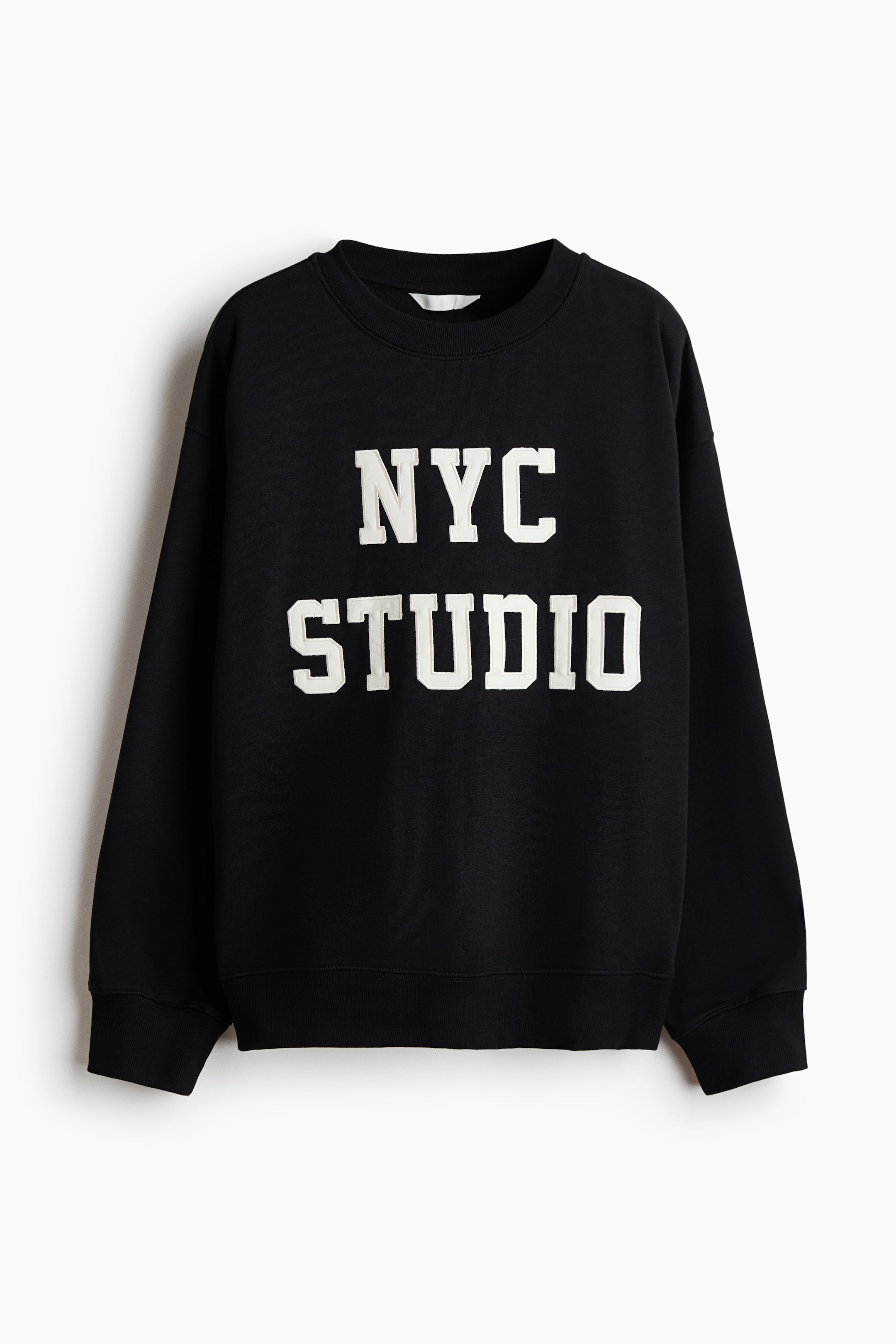 View larger image: An oversized black sweatshirt is hanging facing forward, displaying "NYC STUDIO" in bold, white block letters across the chest. The garment features a ribbed crew neckline, dropped shoulders, long sleeves, and ribbed detailing at the cuffs and hem.