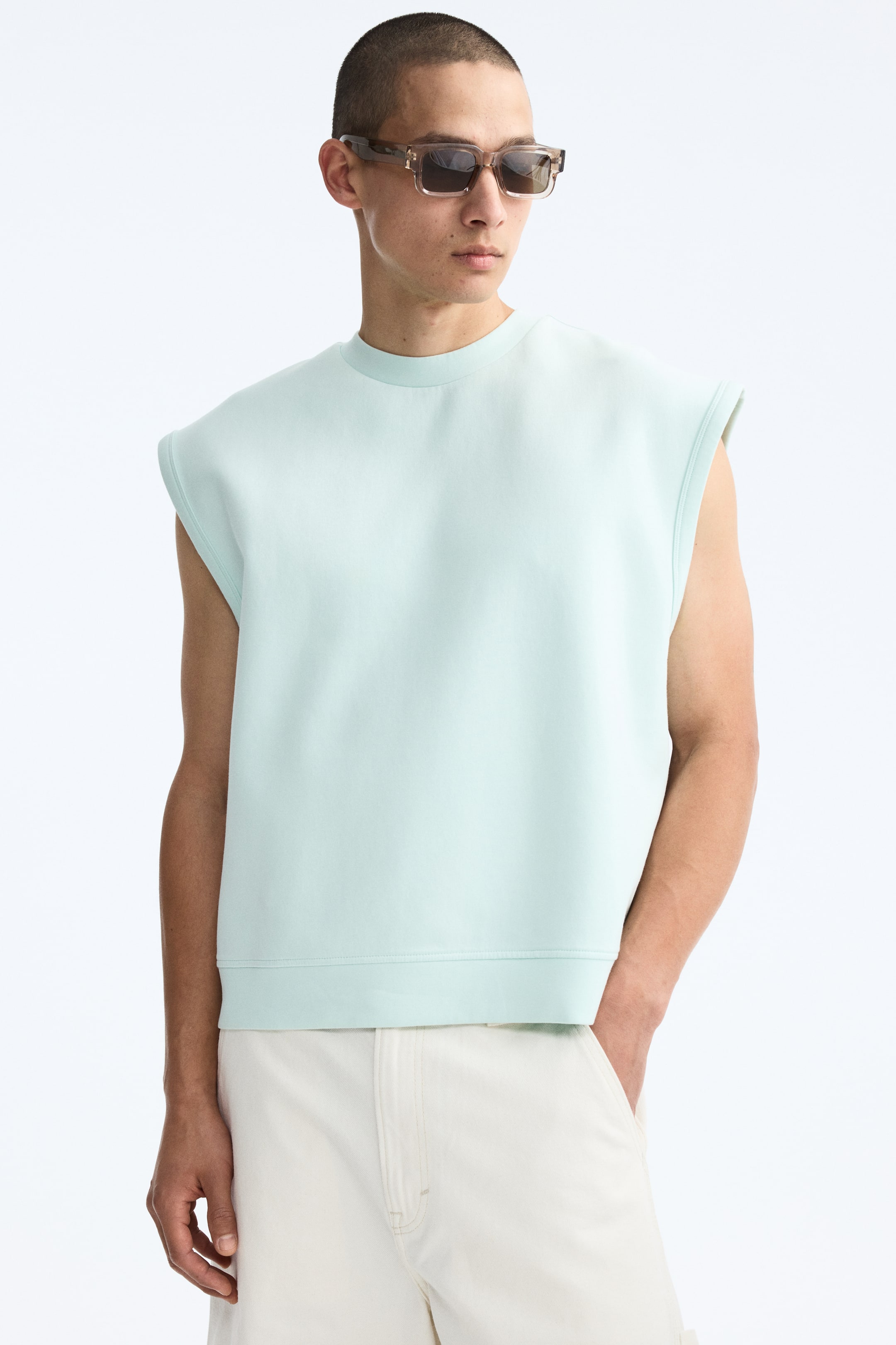 View larger image: The model is wearing a light green tank top, crafted from a smooth, structured fabric, featuring a round neckline, deep armholes, and a straight-cut hem. She pairs it with light-colored shorts, with one hand casually resting in a shorts pocket.