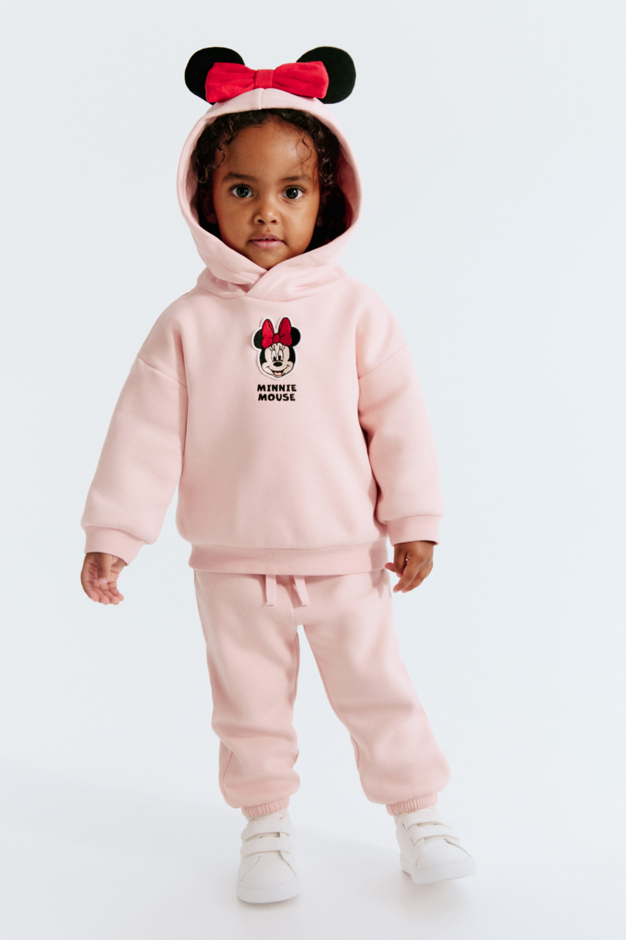 View larger image: A light pink fleece hoodie and track pants set is shown from the front, featuring a hoodie with a Minnie Mouse graphic on the chest and a hood detailed with 3D ears and a red bow. The matching track pants are styled with an elasticated waistband, a decorative drawstring, and elasticated hems.