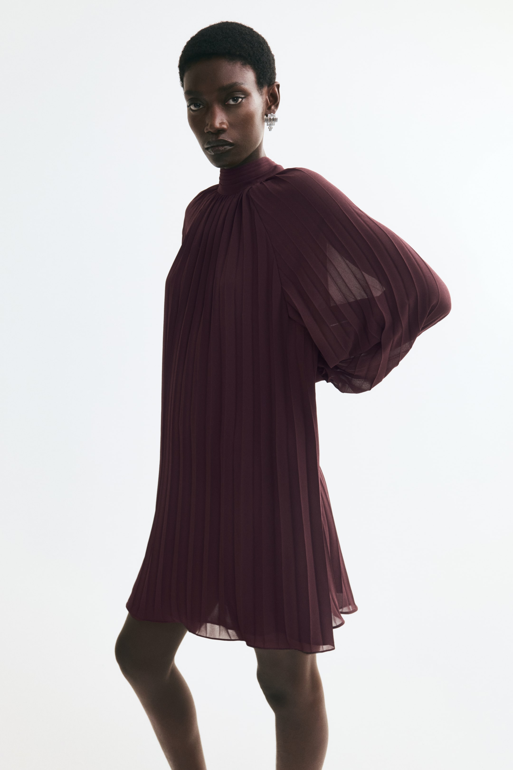 Ver imagen más grande: La modelo lleva un minivestido burdeos plisado con cuello alto y mangas largas y voluminosas. El tejido transparente y ligero cae en una silueta holgada con plisado vertical en toda la prenda.