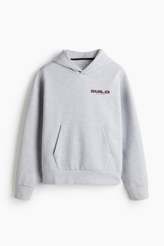 En sportig hoodie med oversized passform i ljusgrått presenteras från FRAMSIDAN, med en korsad huva, en stor känguruficka och ribbade muddar och fåll. En mörkröd "BUILD"-grafik är synlig på bröstet, tillsammans med en "DryMove™" och "Oversized Fit"-etikett inuti halsringningen. - 1