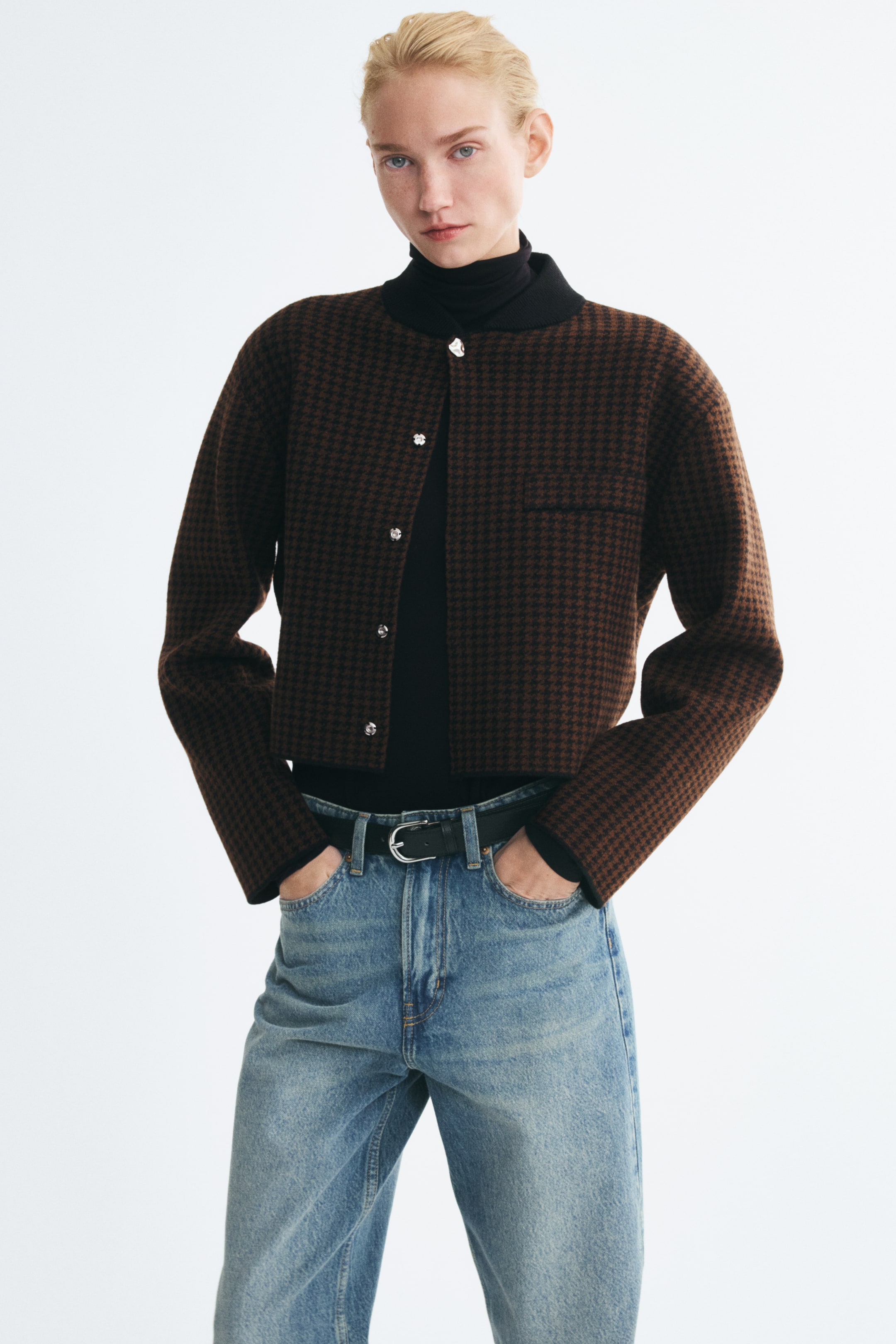 Ampliar la imagen: La modelo lleva un cárdigan de punto corto con estampado de pata de gallo sobre un suéter de cuello alto negro y jeans azules. El cárdigan presenta un cuello acanalado negro, un bolsillo de ribete en el pecho y botones color plata, mientras la modelo posa con las manos metidas en los bolsillos de los jeans.
