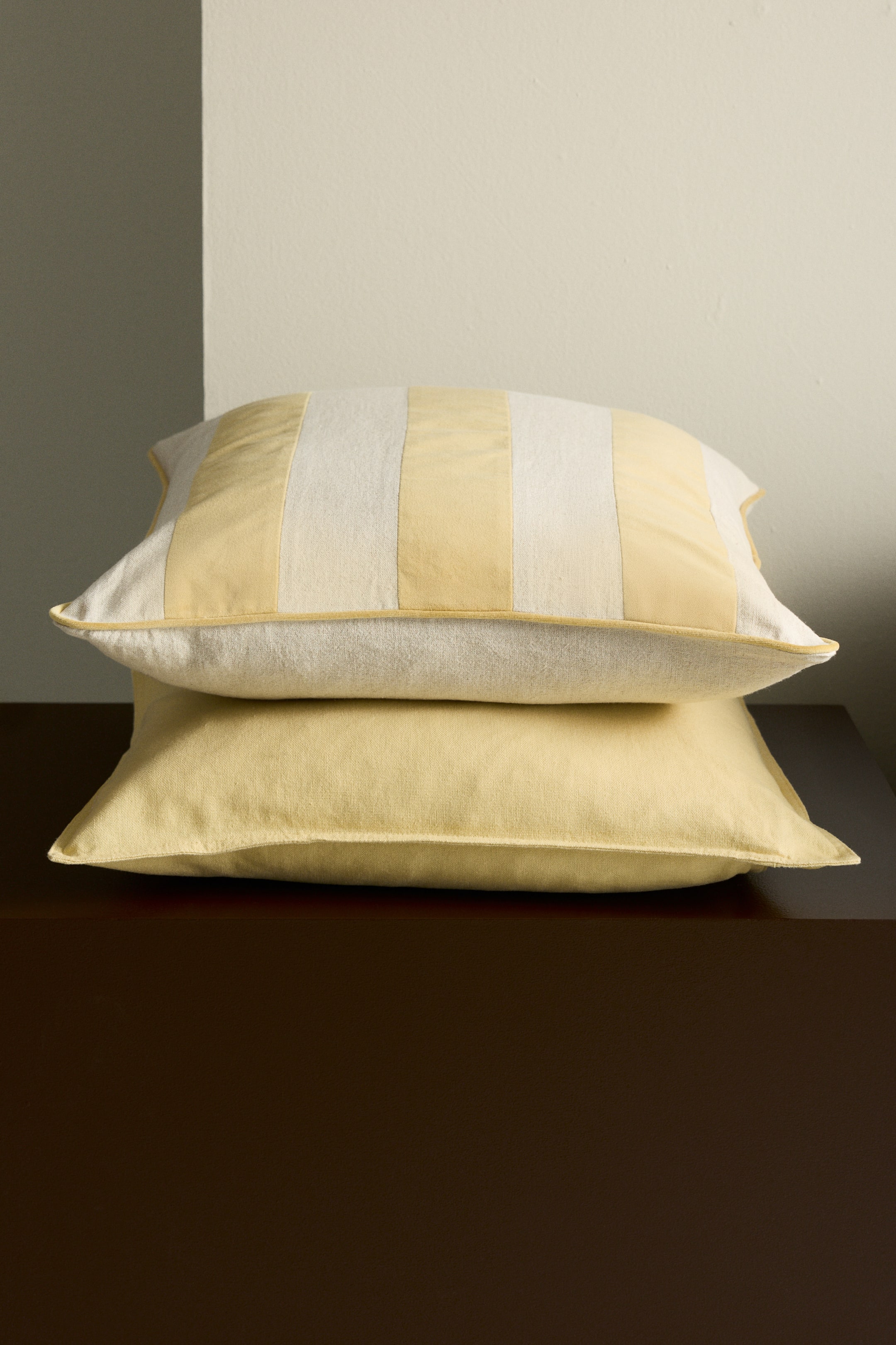 Visualizza immagine più grande: Due cuscini rettangolari sono impilati, presentati con un leggero angolo. Il cuscino inferiore è a tinta unita di un giallo chiaro; il cuscino superiore presenta righe verticali in giallo chiaro e avorio, con piping giallo chiaro che ne definisce i bordi.