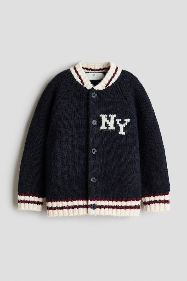 A loose-fit cardigan in a dark navy blue soft knit is shown facing front, featuring a white "NY" motif on the chest and a button-down closure. The design includes long raglan sleeves, a small ribbed collar, and matching ribbed cuffs and hem in off-white with burgundy striped accents. - 1